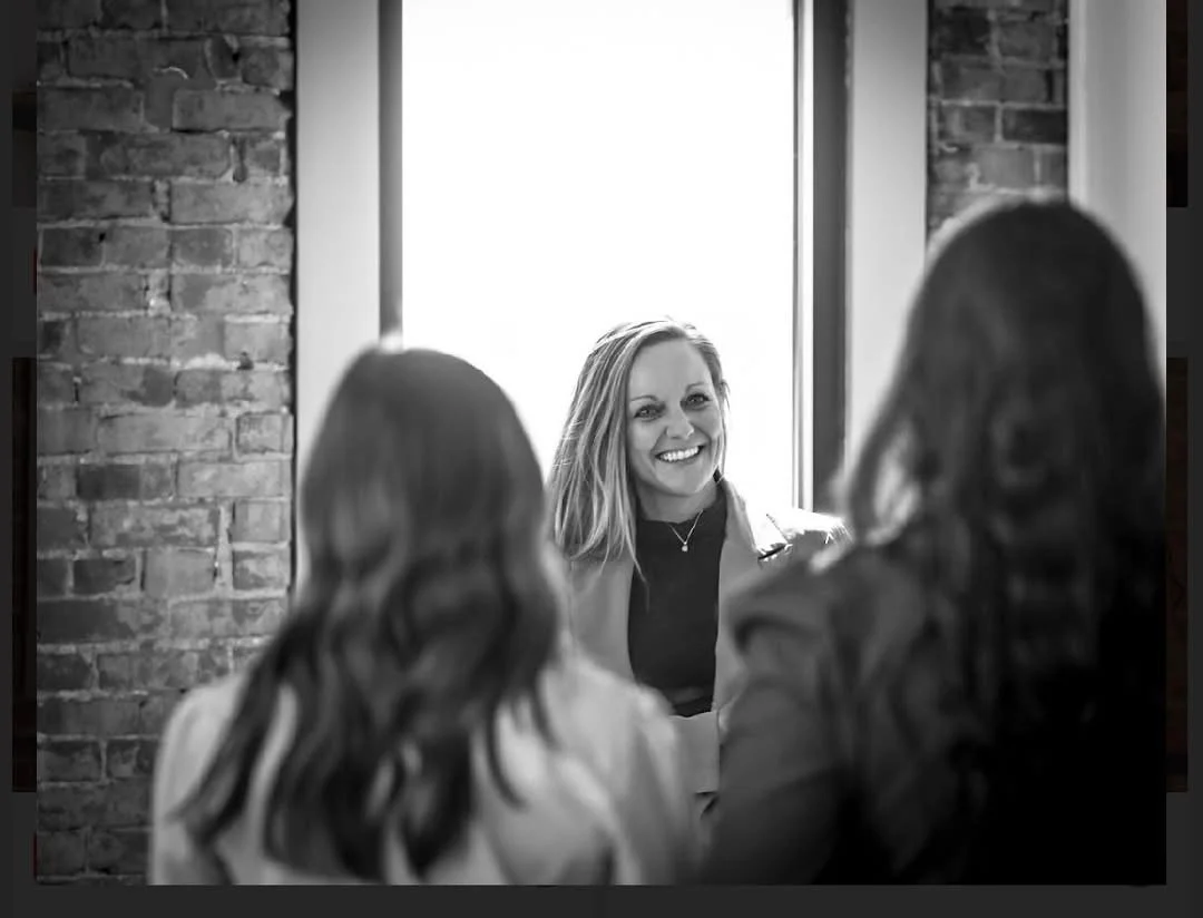 A woman smiling while talking to two other women in front of a window with brick walls on either side.