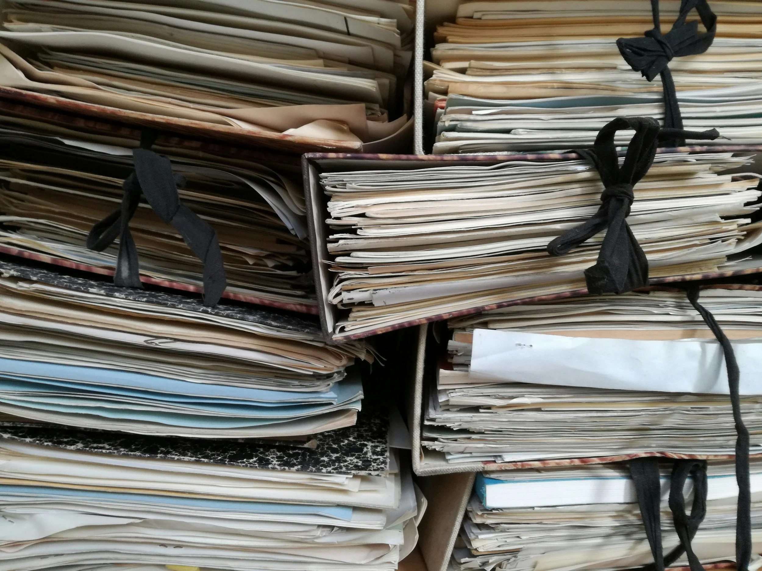Stacks of disorganized papers and files in cardboard boxes tied with black fabric strips.