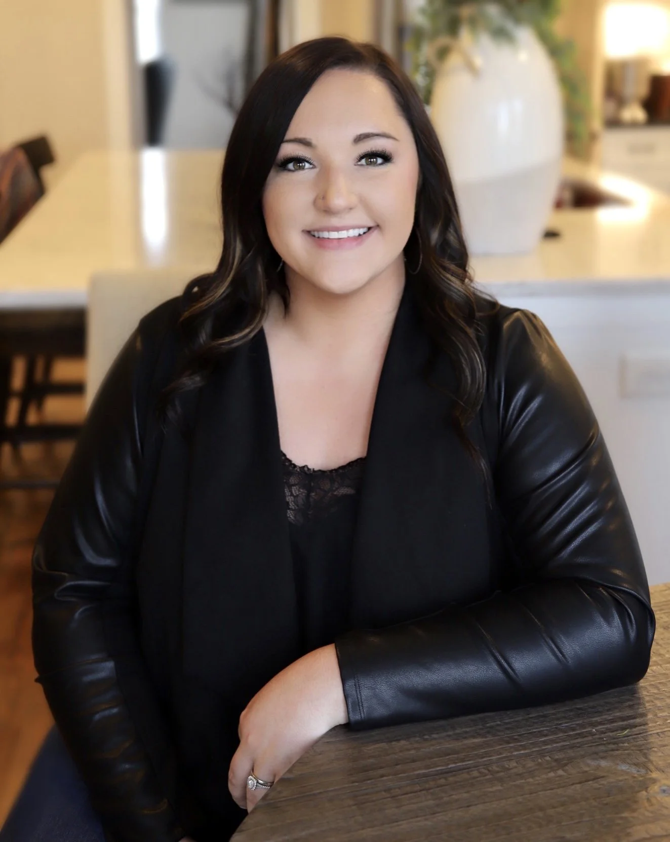 A woman with dark hair, wearing a black leather jacket and lace top, sitting at a wooden table in a well-lit home interior, smiling at the camera.