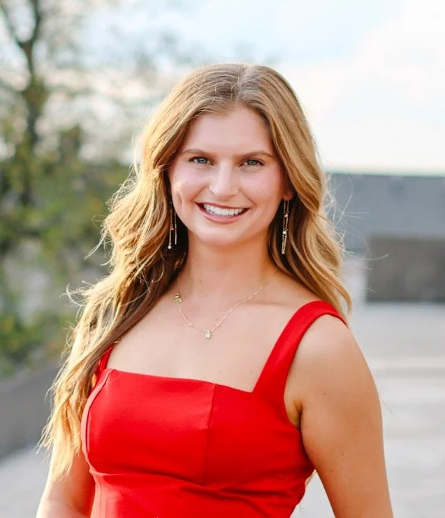 Young woman with long blonde hair wearing a red sleeveless dress, smiling outdoors.