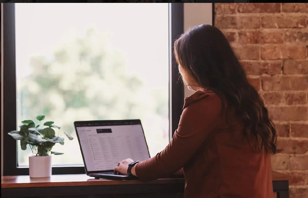 A woman with long dark hair in a brown top working on a laptop at a wooden desk by a window with a potted plant nearby.