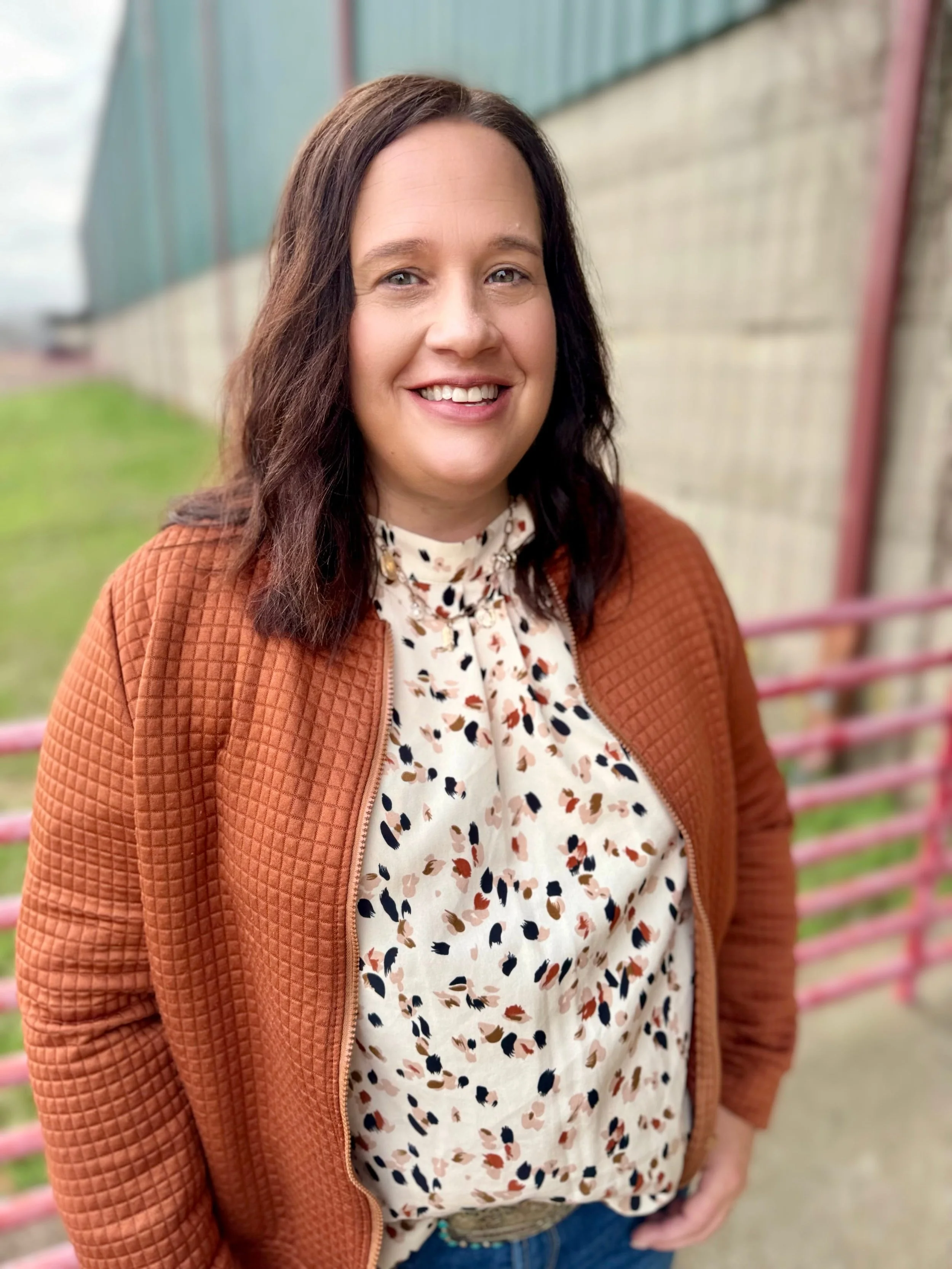 A smiling woman with shoulder-length dark hair outdoors, wearing a brown quilted jacket and a cream blouse with a multicolored animal print, standing in front of a brick wall and a pink metal railing.