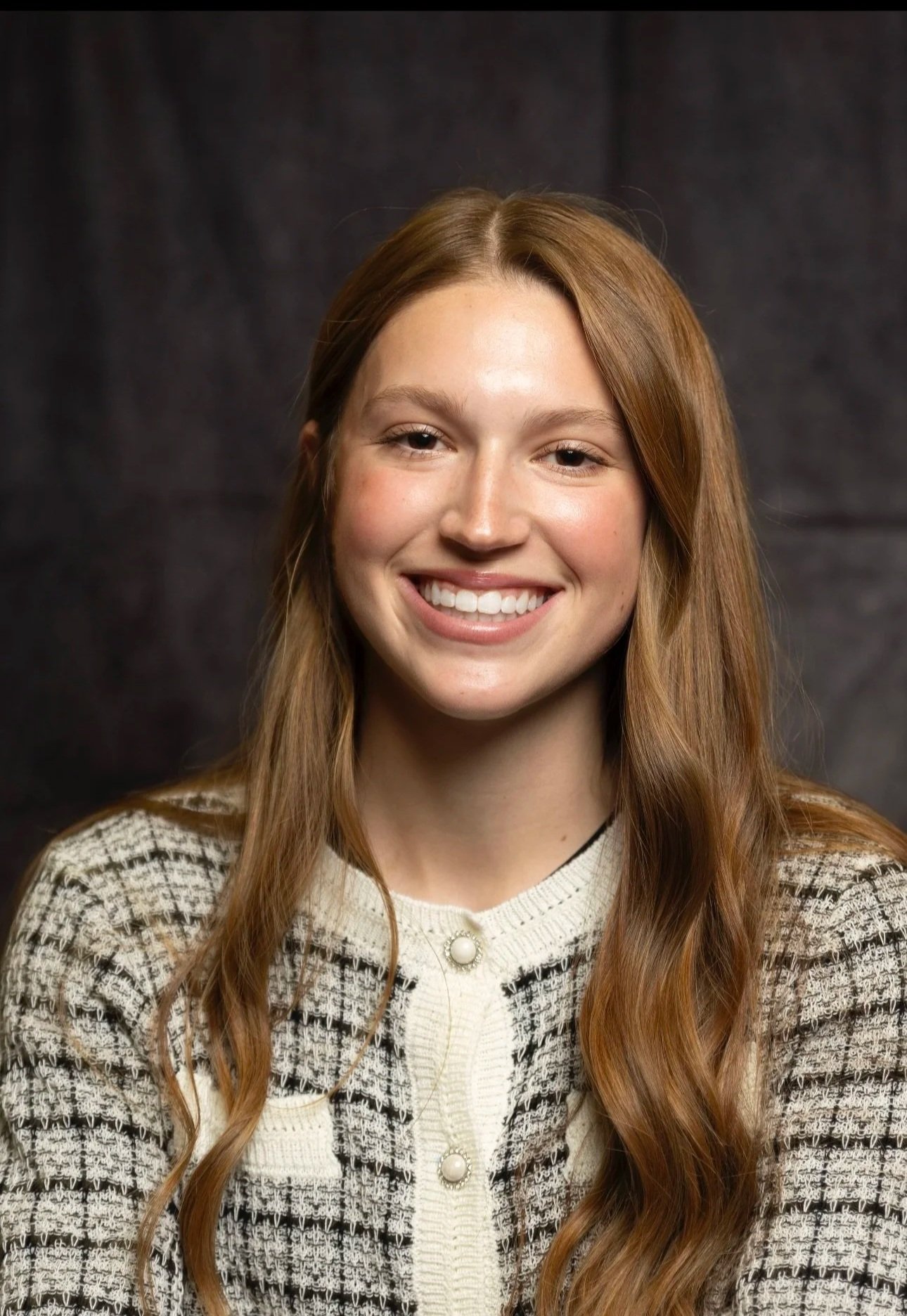 A young woman with long, wavy, red hair smiling at the camera. She is wearing a cream-colored, buttoned cardigan with black accents and a pearl detail near the collar, against a dark, textured background.
