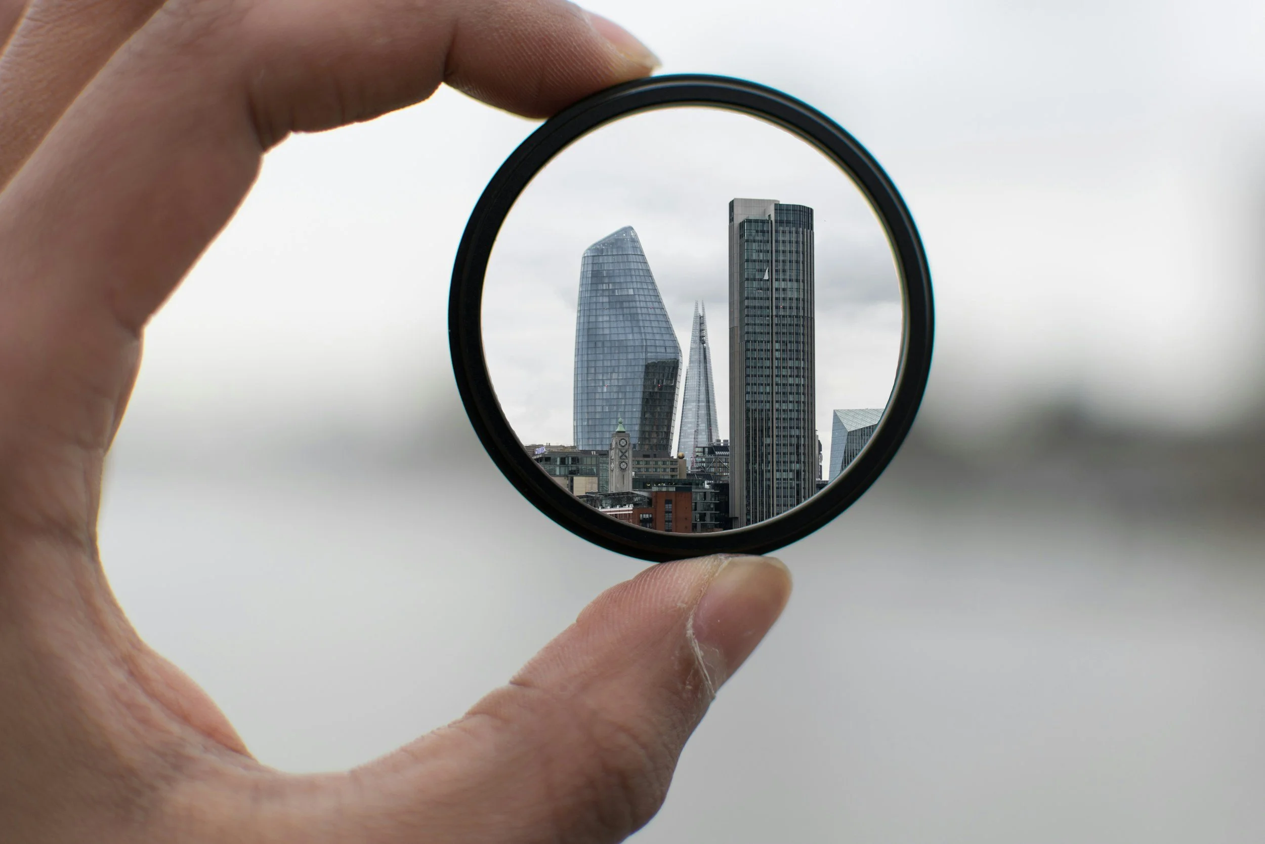 A person's hand holding a circular lens or filter, showing a clear view of the London skyline, including the Shard, the Walkie Talkie building, and other skyscrapers, against a cloudy sky.