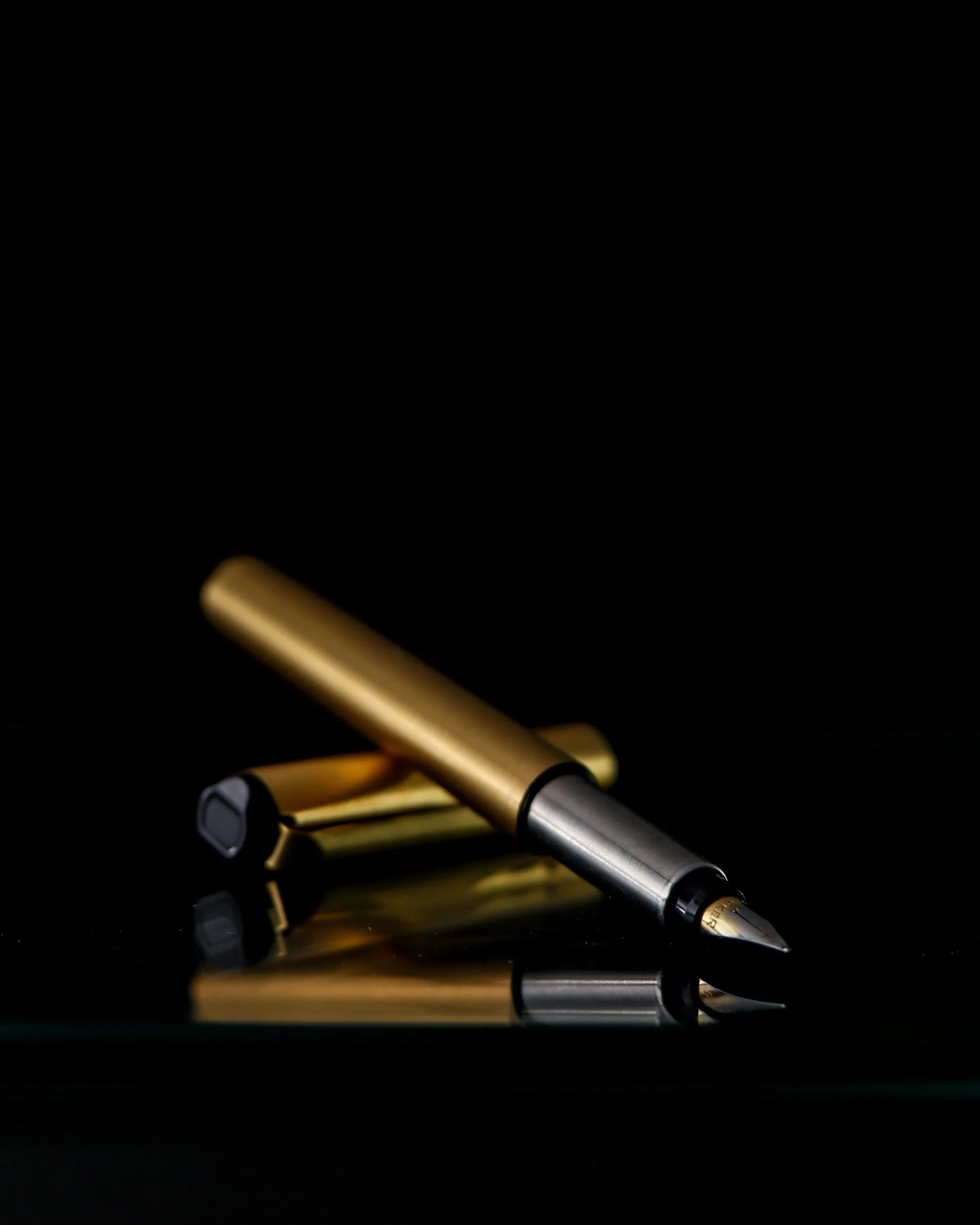 Close-up of a gold and black fountain pen with a silver nib resting on a reflective black surface against a black background.