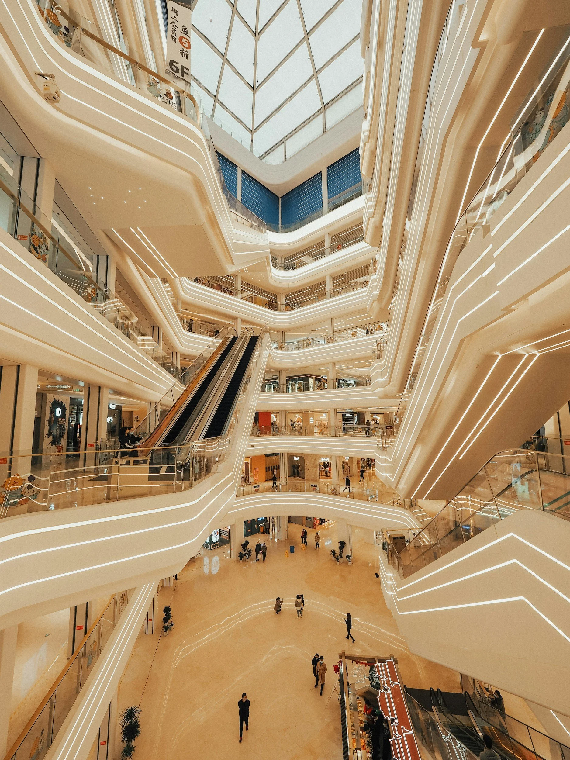 Interior view of a modern multi-level shopping mall with curved white balconies, glass railings, escalators, and bright lighting.