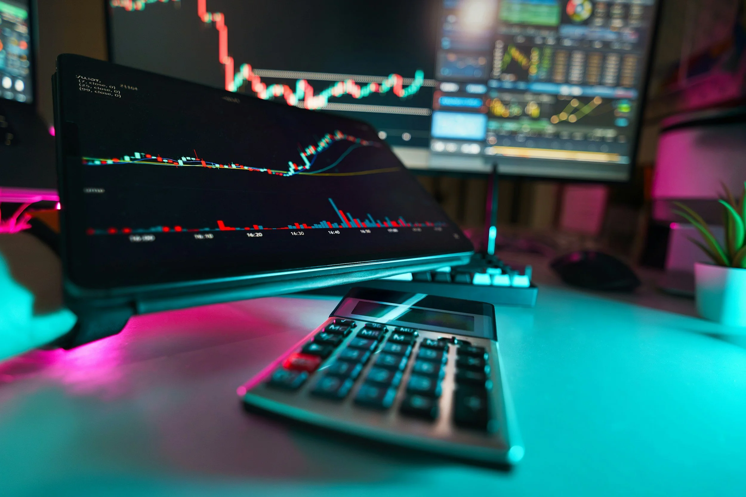 Multiple computer screens displaying stock trading charts, a calculator on the desk, and a potted plant, illuminated by colorful LED lighting.