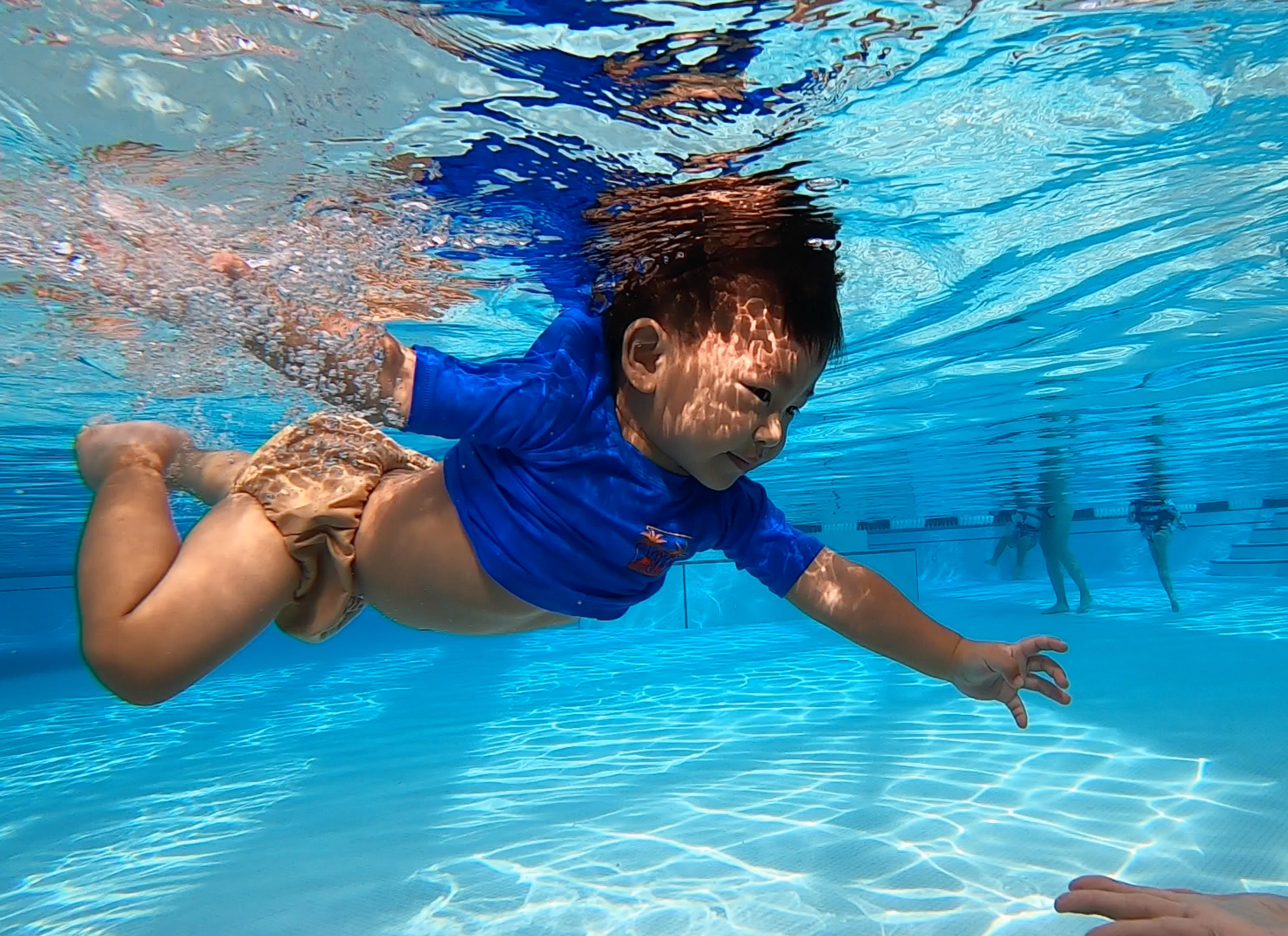 A young boy swimming underwater in a pool, reaching out with one hand, wearing a blue shirt and tan swim shorts.