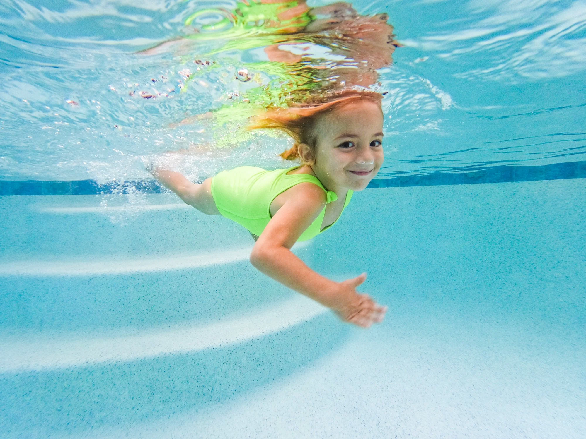 A young girl in a bright green swimsuit swimming underwater and smiling at the camera in a swimming pool.
