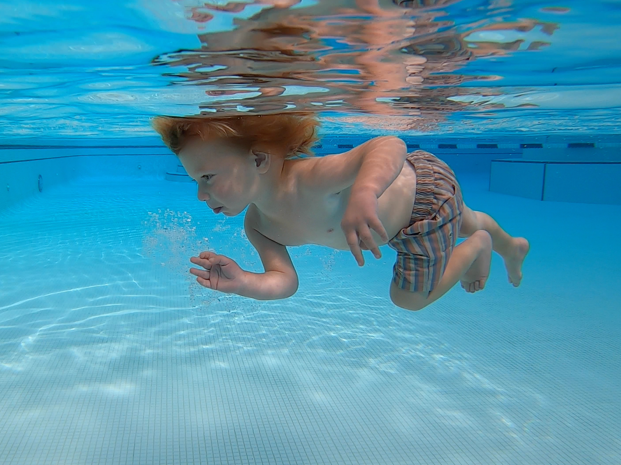 Young boy swimming underwater in a pool, wearing striped swim trunks.