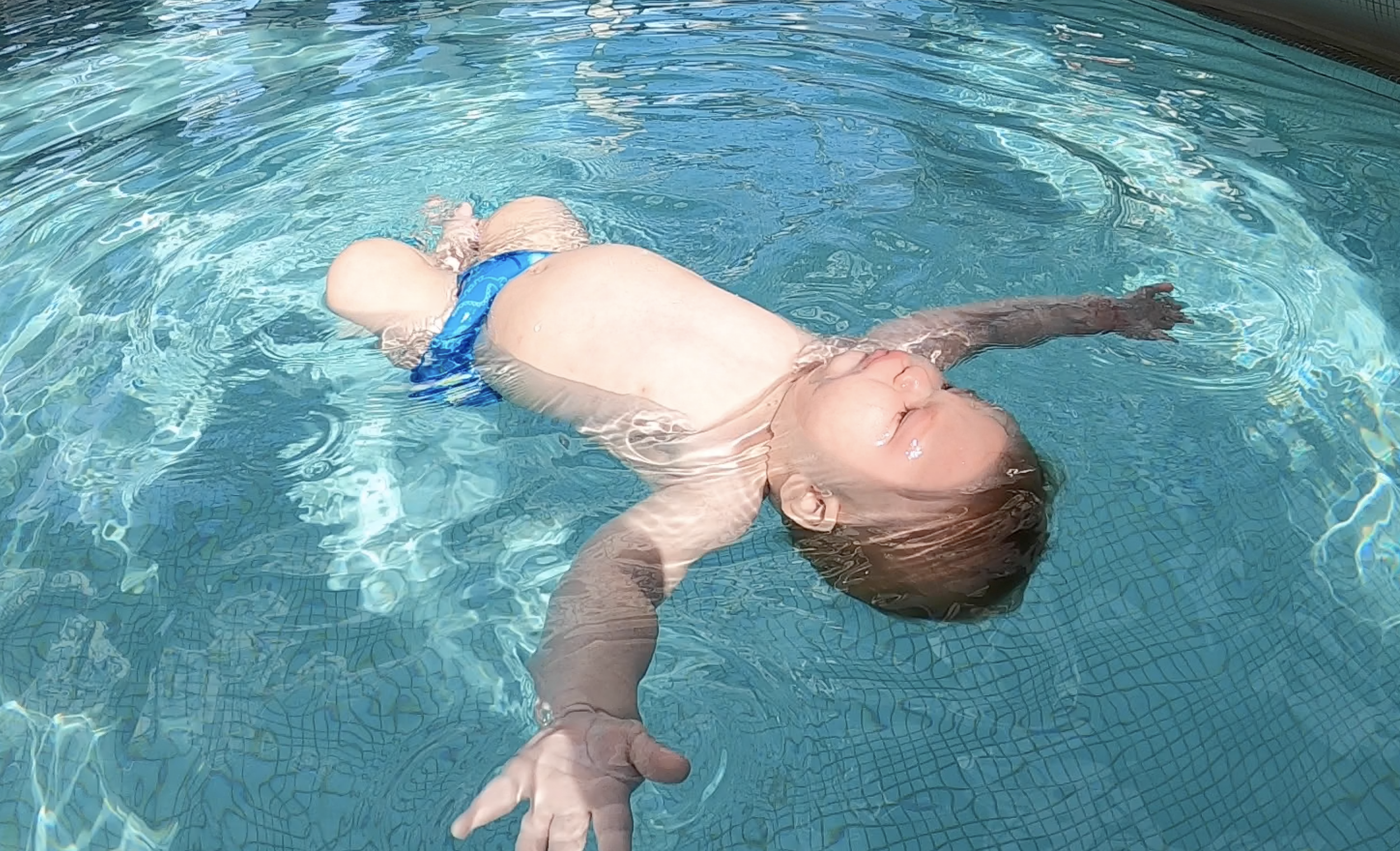 A young child floating on their back in a swimming pool, wearing blue swim trunks, with their eyes closed and their arms extended outward.