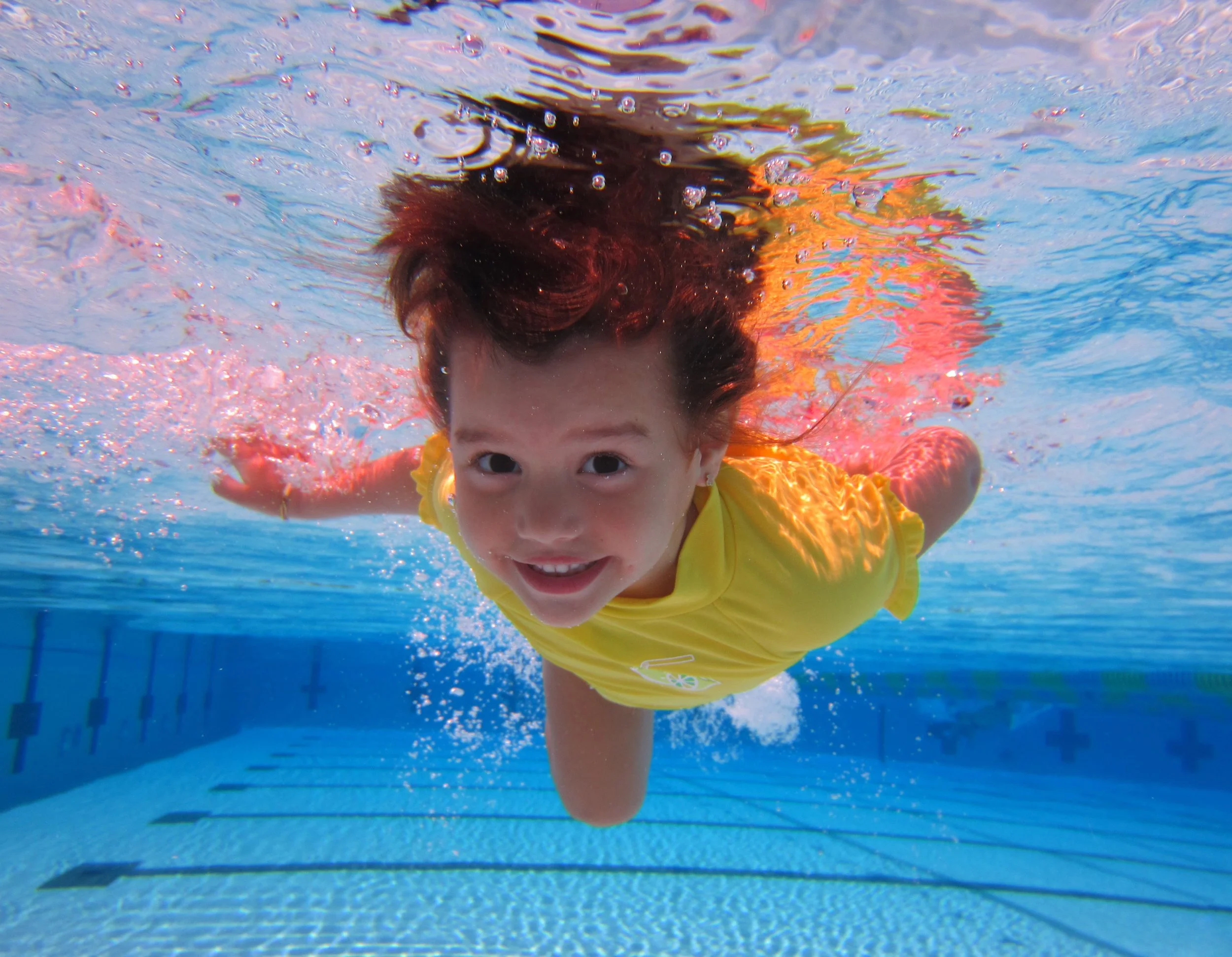 Young child with brown hair swimming underwater in a pool, wearing a yellow shirt, looking at the camera, with blue water and lane markings in the background.