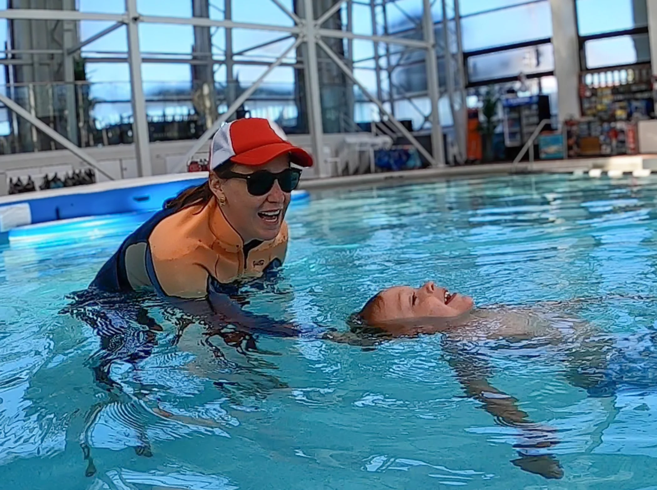 A woman in sunglasses and a red and white cap assists a smiling boy with brown hair in a swimming pool.