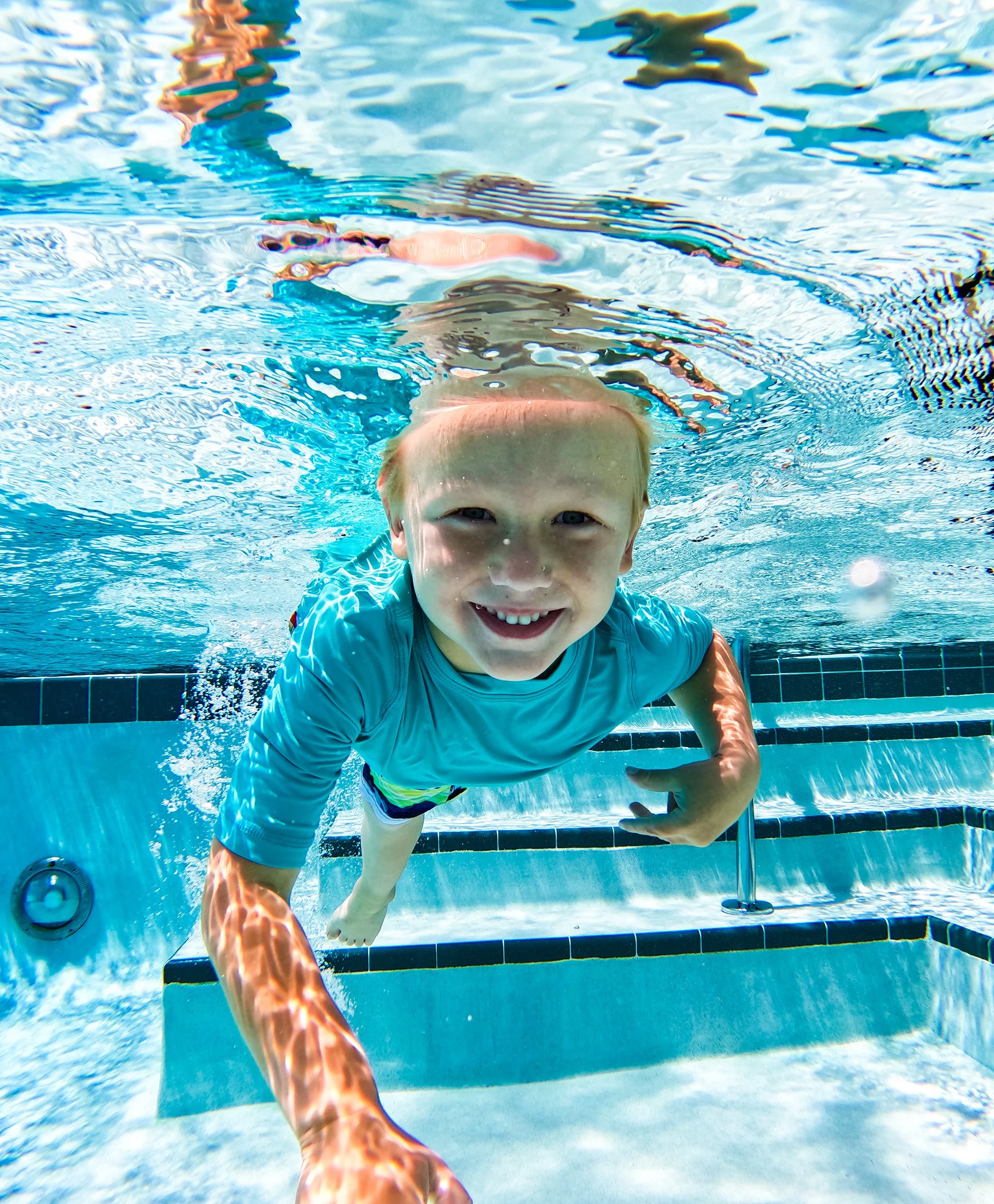 Smiling young boy swimming underwater in a pool, wearing a blue shirt.