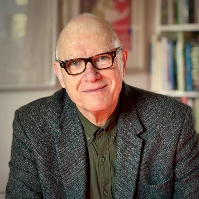 An older man with glasses, wearing a dark green shirt and a gray blazer, smiling in front of bookshelves.
