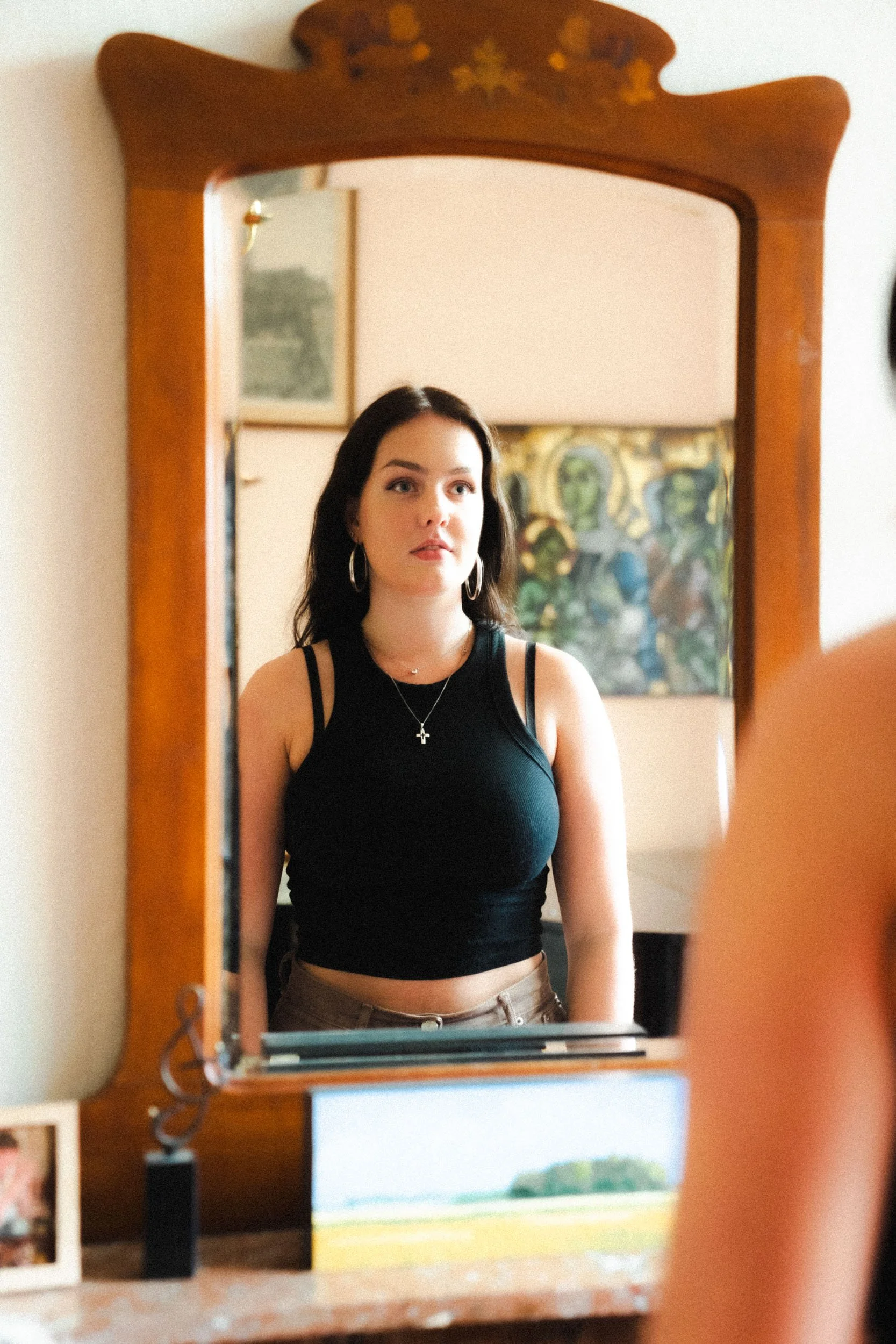 A young woman with dark hair and hoop earrings looking at her reflection in a mirror. She is wearing a black tank top and brown pants, with a cross necklace.
