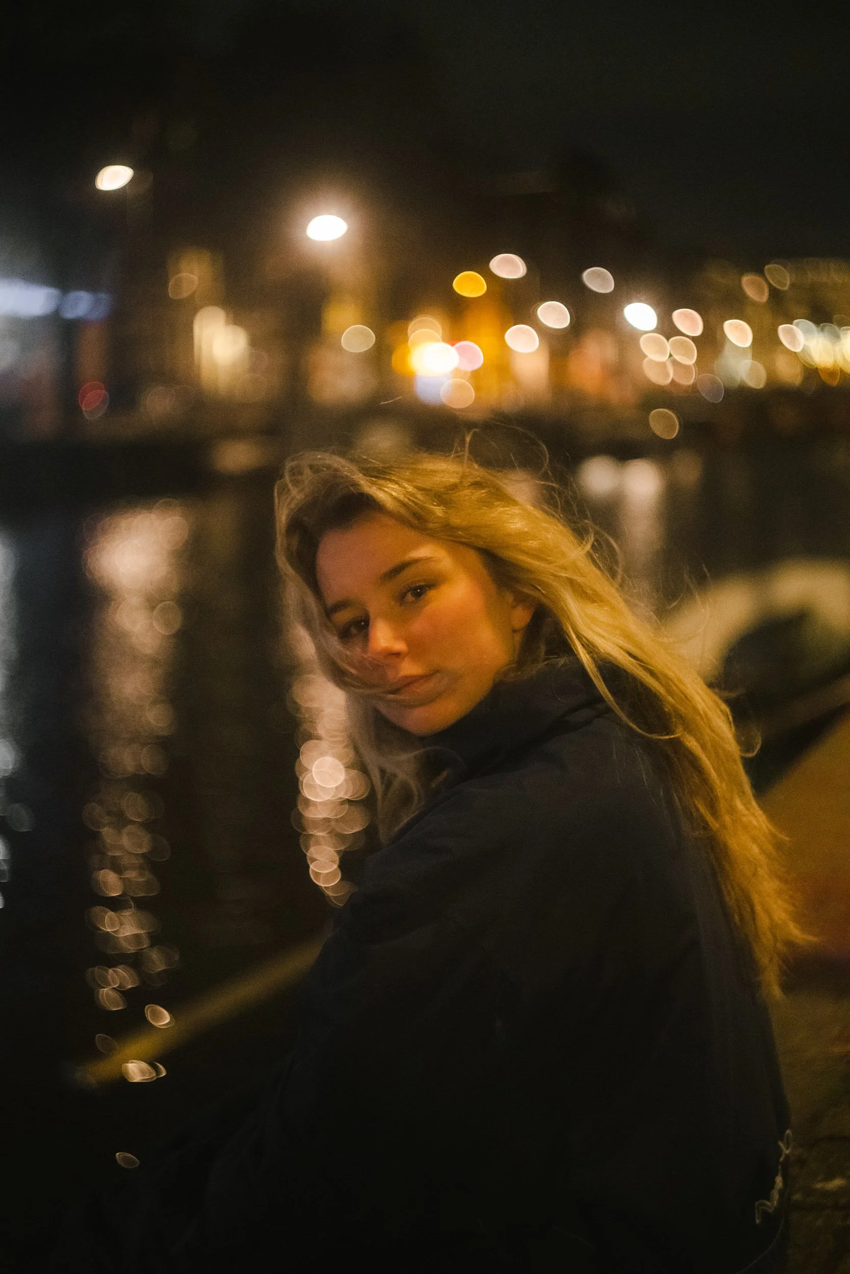 A young woman with blonde hair sitting by a river at night with blurred city lights in the background.