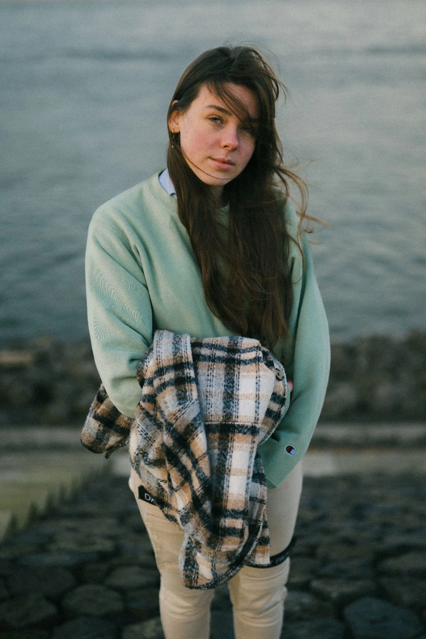 A young woman with long brown hair and jewelry standing outdoors near water, holding a plaid jacket, wearing a light green sweatshirt and beige pants.