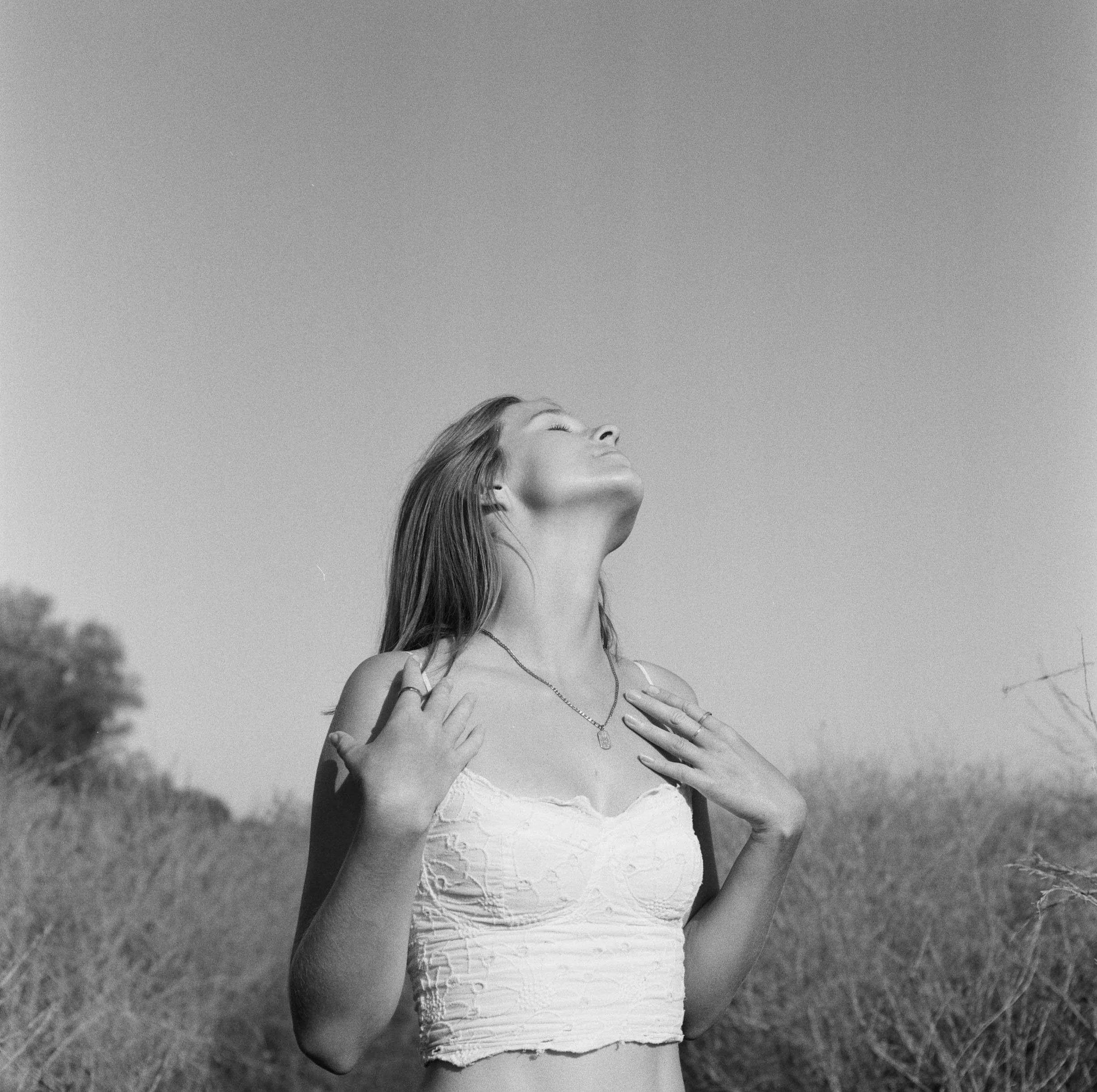 A woman standing outdoors on a grassy field with eyes closed, head tilted slightly upward, wearing a sleeveless top and a necklace.