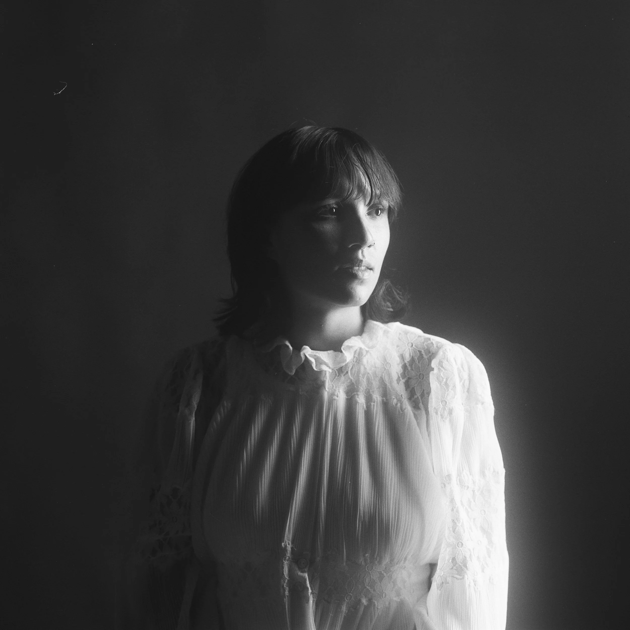 A black and white portrait of a woman with shoulder-length hair, wearing a lace and pleated blouse, looking to her left with a soft expression.