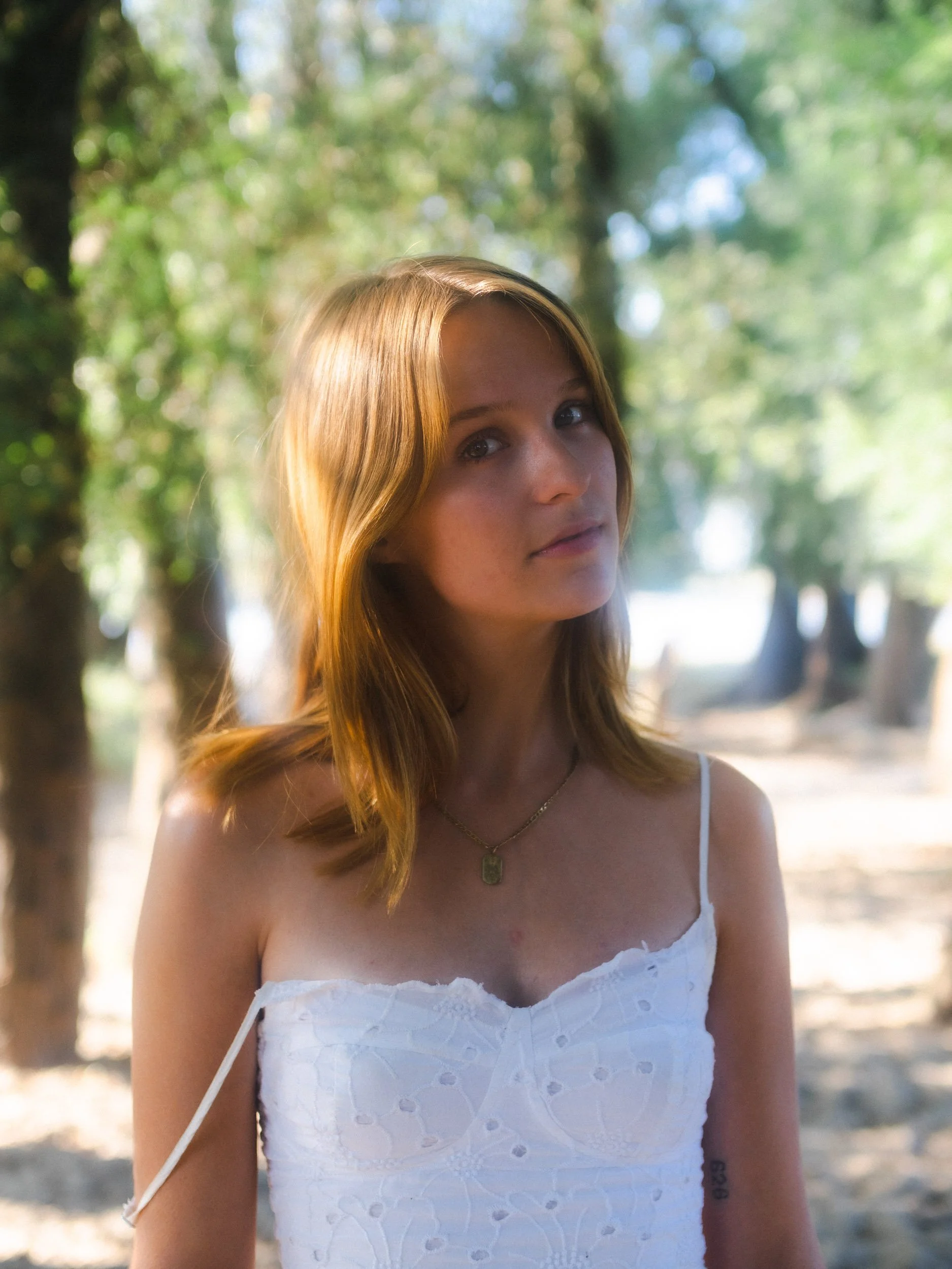 Young woman with reddish hair wearing a white tank top and a gold necklace outdoors in a wooded area.