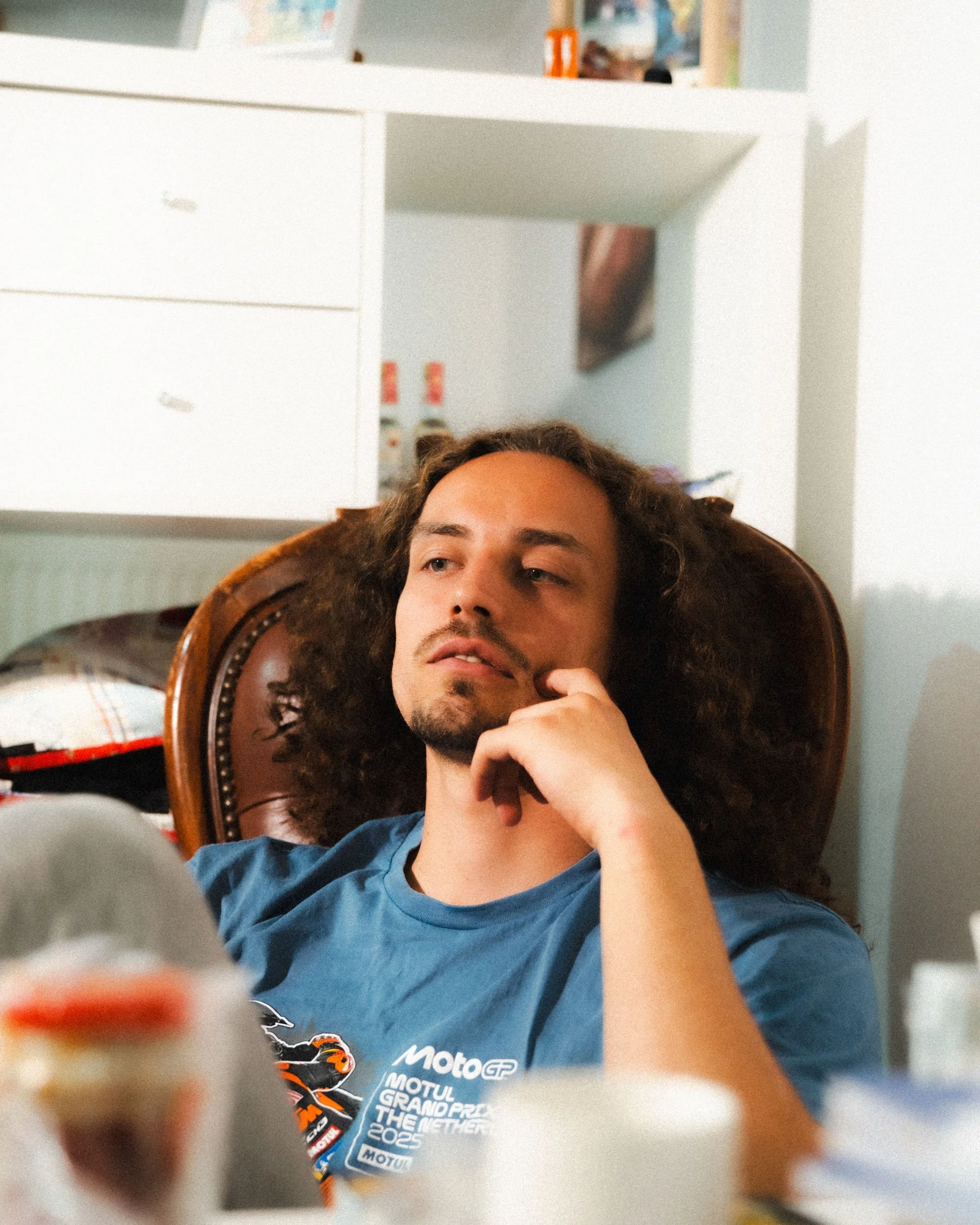 A man with long curly hair and a goatee sitting on a brown leather chair, resting his chin on his hand, wearing a blue MotoGP T-shirt. In the background, there is a room with white shelves, some small containers, and a poster on the wall.