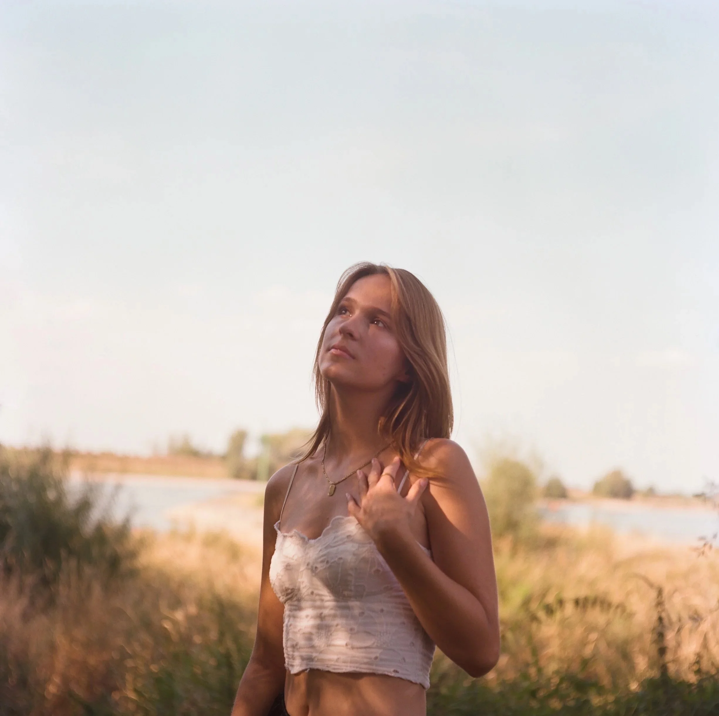 A young woman with shoulder-length brown hair wearing a light-colored crop top and necklace stands outdoors near water, gazing thoughtfully into the distance.