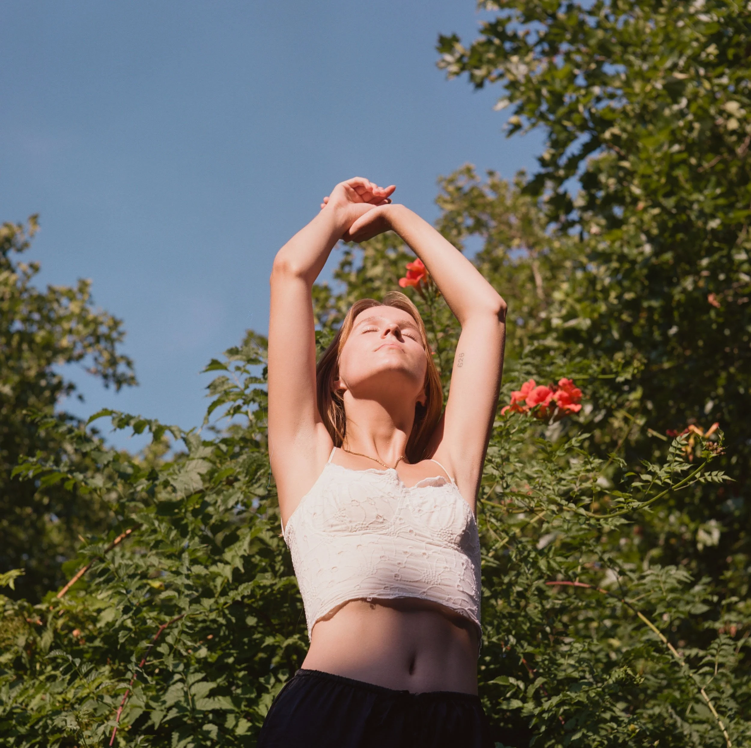 A woman with red hair stands outdoors with her eyes closed and arms raised above her head, basking in sunlight amidst green foliage and pink flowers.