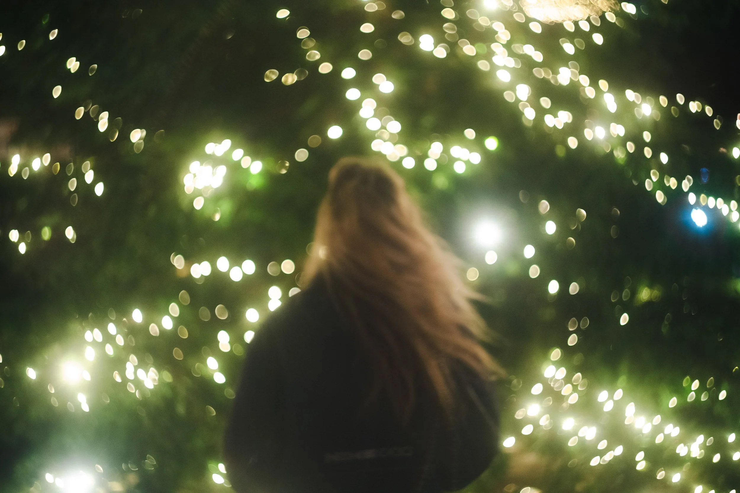 A person with long hair, seen from behind, standing in front of a tree decorated with fairy lights at night.