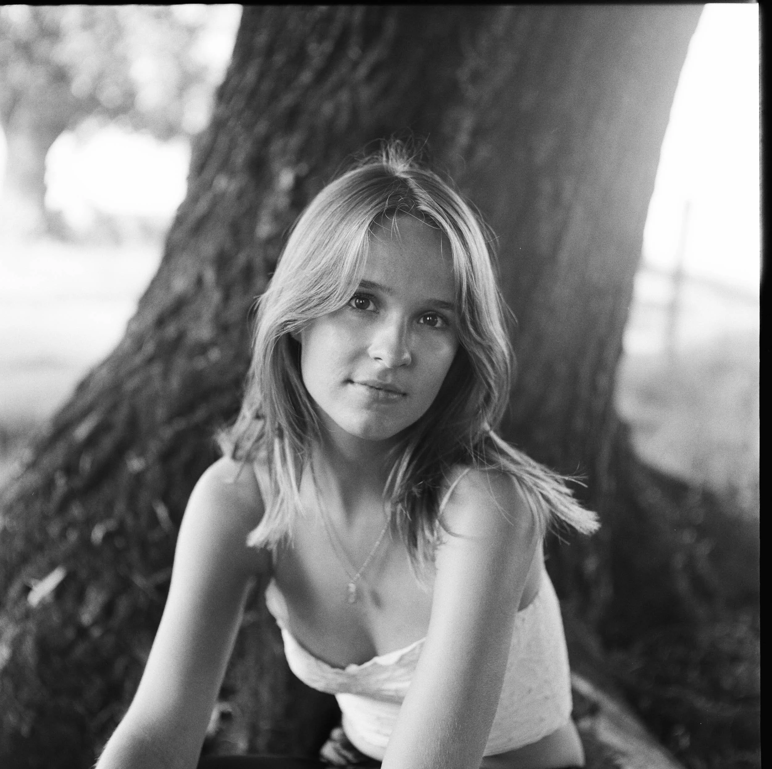 A young woman with shoulder-length hair sitting outdoors next to a large tree, looking at the camera with a slight smile. The photo is in black and white.
