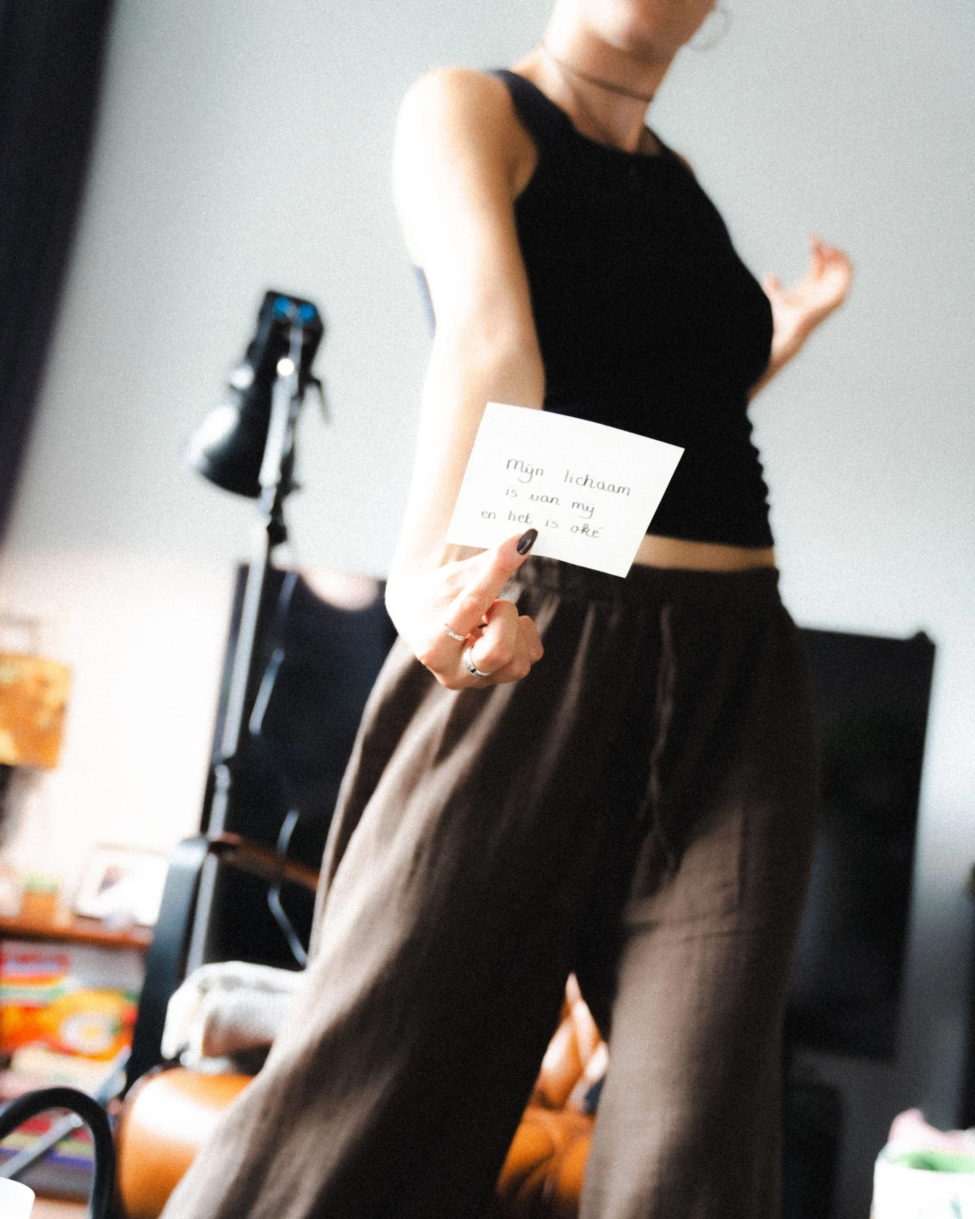 A woman holding a handwritten note in her right hand, wearing a black sleeveless top and brown pants, standing indoors with a bookshelf, TV, and lamp in the background.
