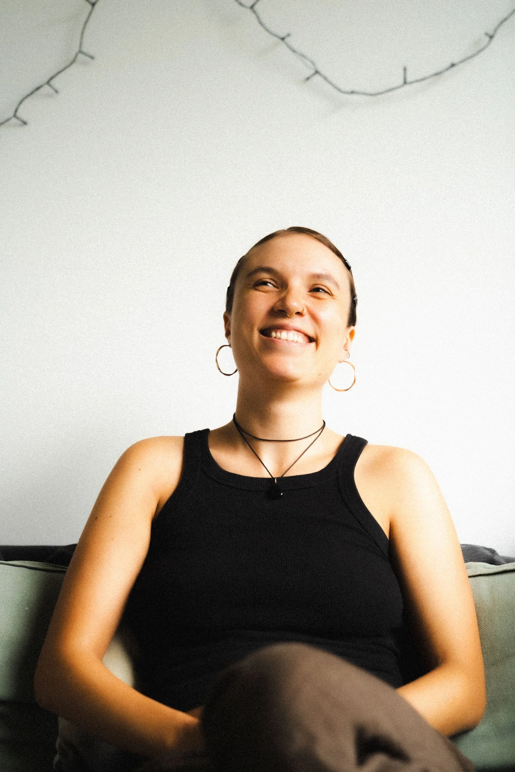 A young woman with short hair smiling while sitting on a sofa, wearing a black tank top, hoop earrings, a choker, and layered necklaces.