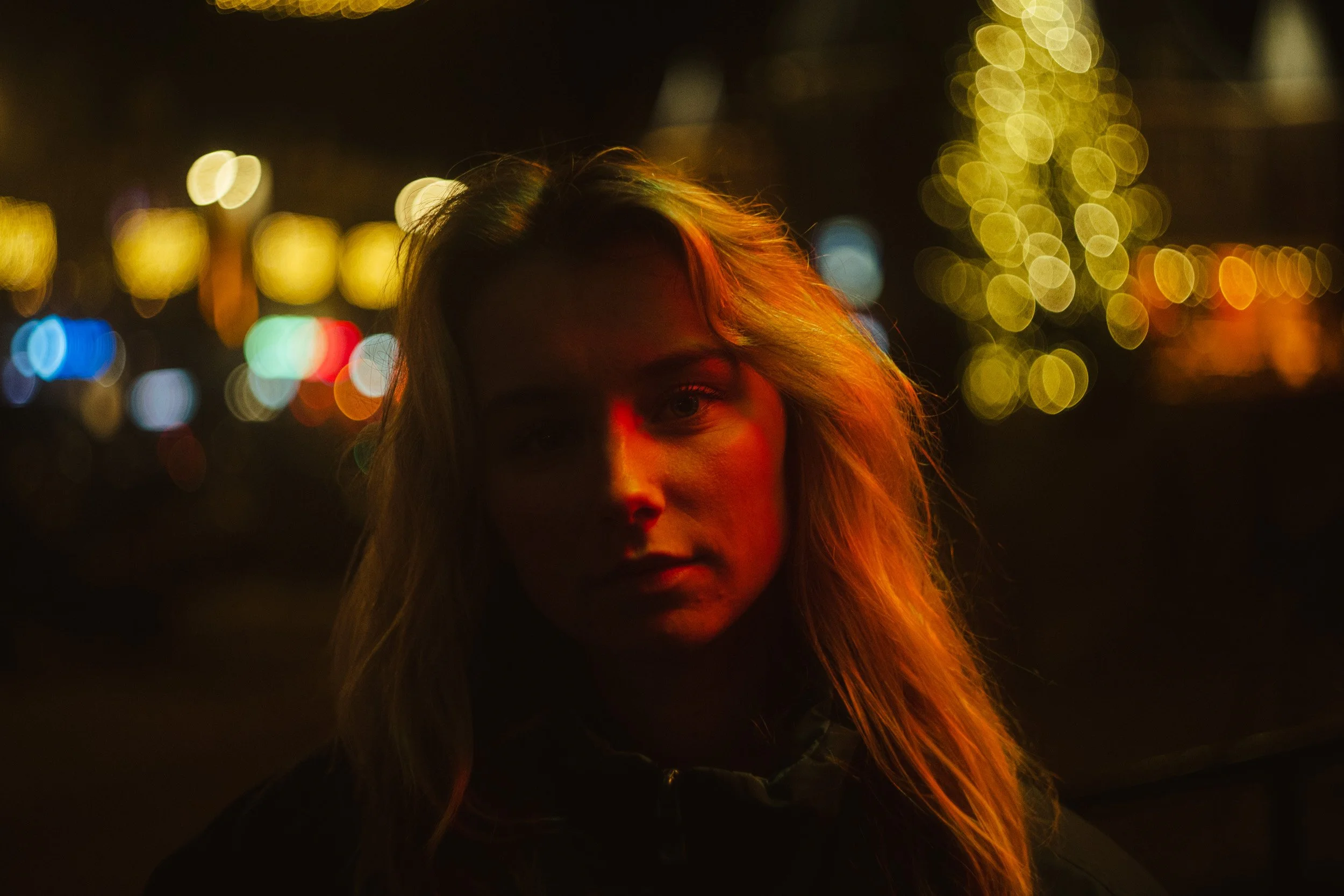 A woman with long blonde hair standing at night with city lights blurred in the background, illuminated by red and orange lighting.