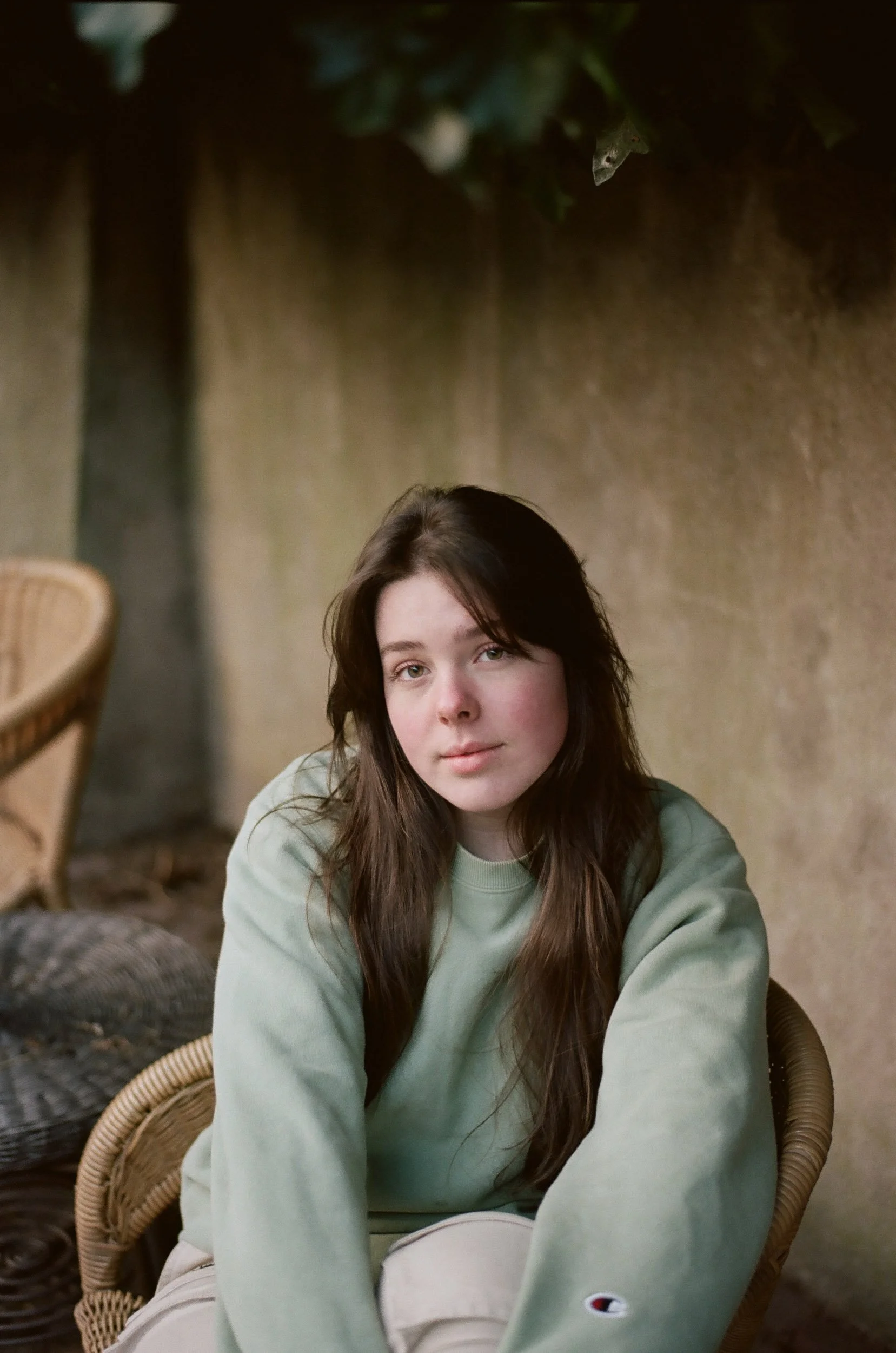 Portrait of a young woman with long brown hair, sitting outdoors against a textured wall, wearing a light green sweatshirt and beige pants, looking at the camera with a neutral expression.