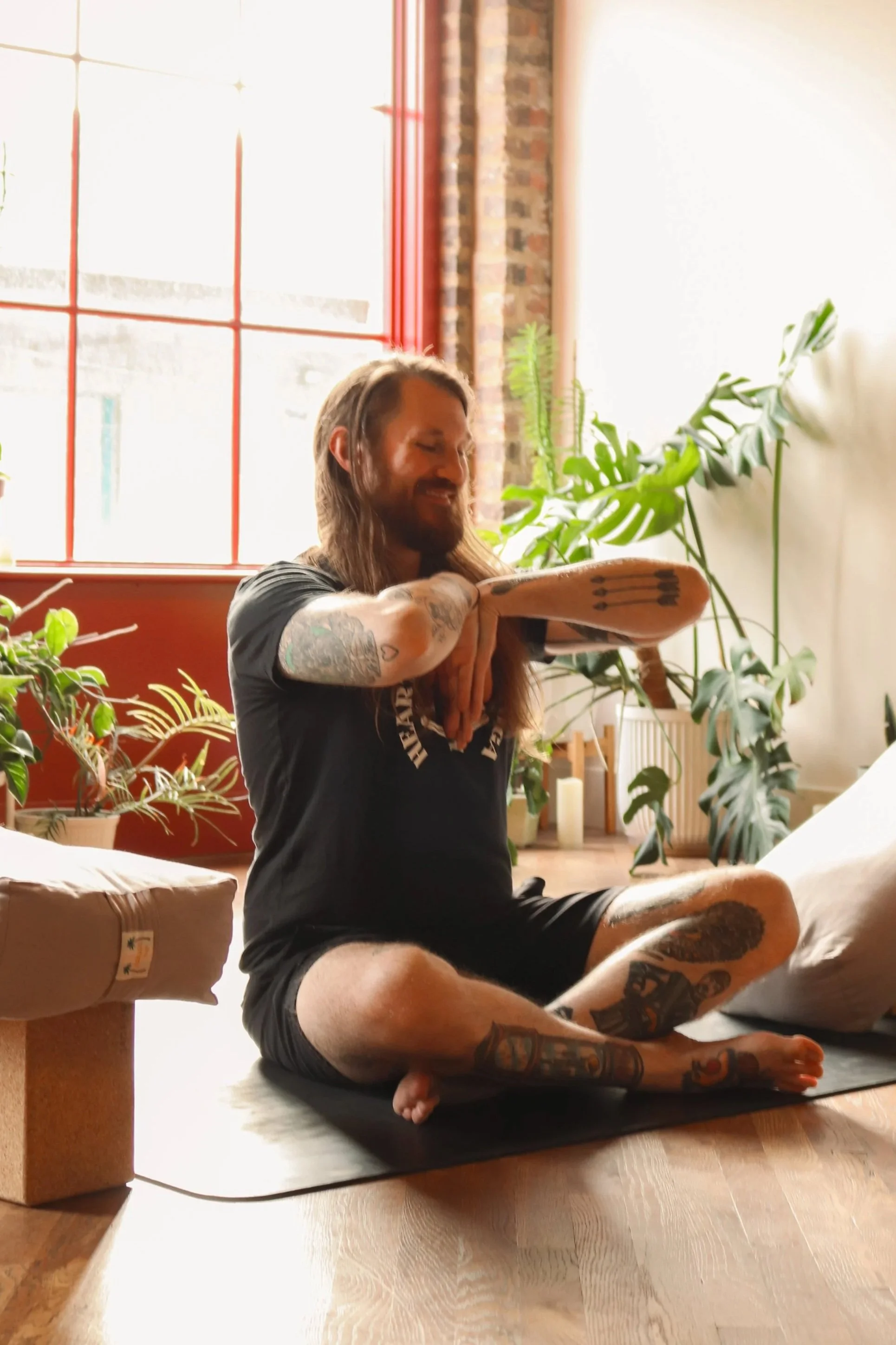 A man with long hair and tattoos on his arms and legs practicing yoga on a black mat in a sunlit room with green plants, large windows, and a brick wall.