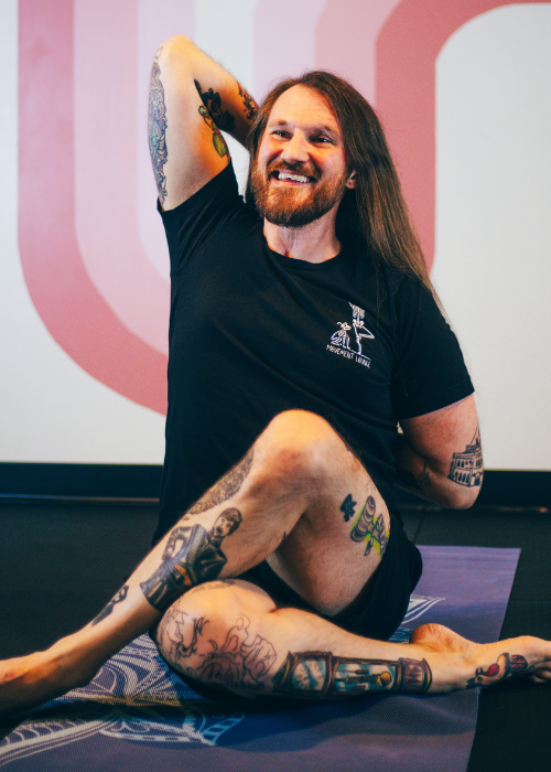 A smiling man with long hair and tattoos practicing yoga on a mat.