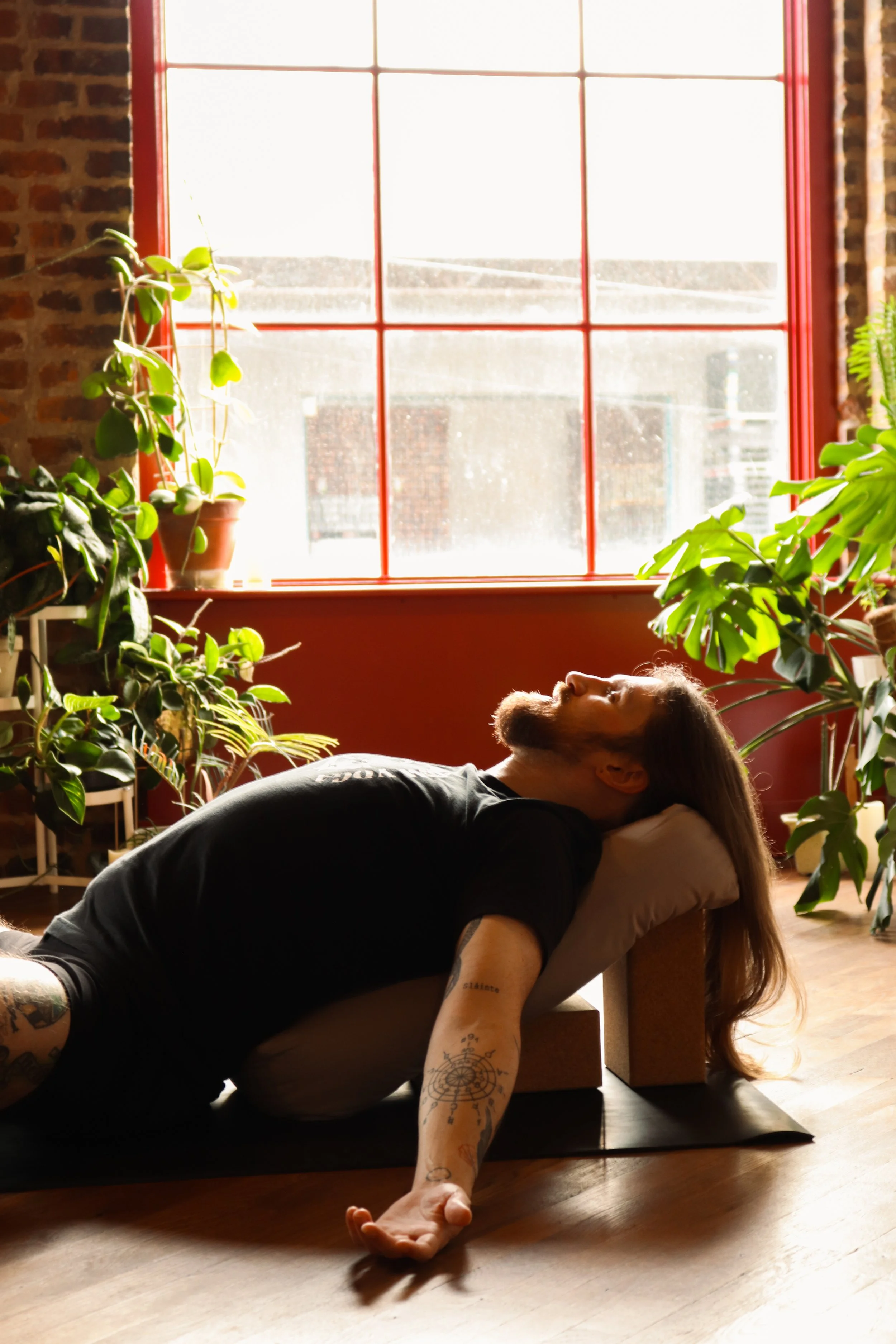 A person lying on a yoga mat doing a stretch with one arm extended and the other arm supporting their head, surrounded by potted plants near a large window with red trim.