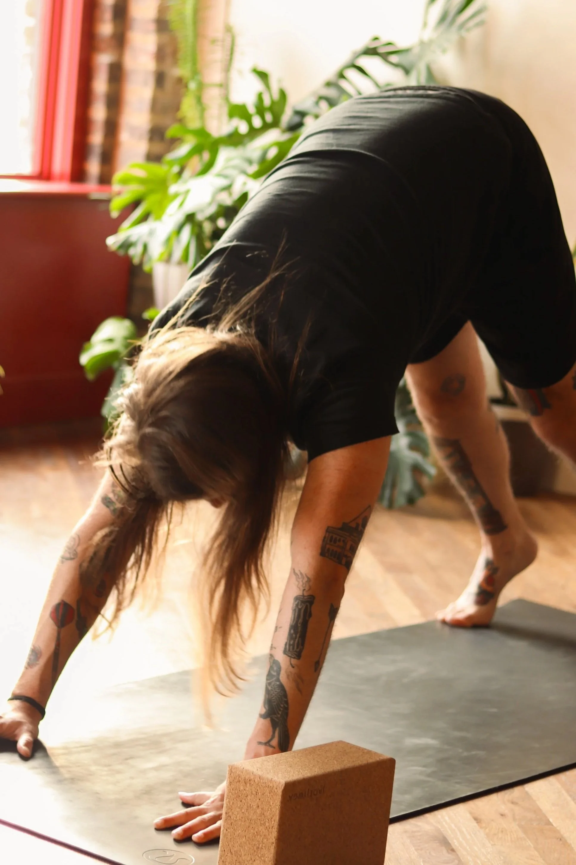 Person with long hair and tattoos in downward dog yoga pose on a mat in a room with plants and a brick wall.