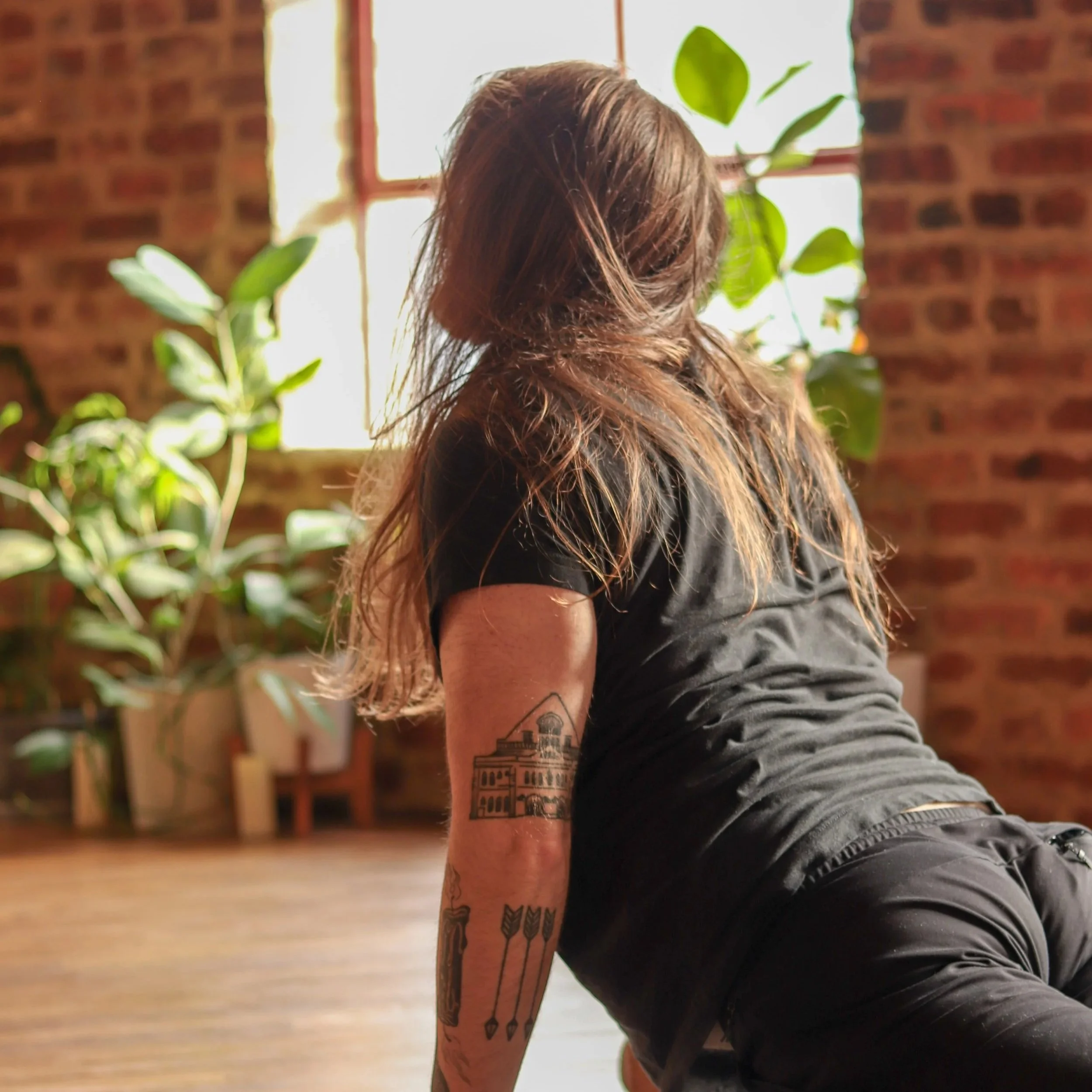 A woman with long, wavy brown hair and tattoos on her arm, wearing a black shirt, is practicing a yoga pose on a wooden floor with a brick wall and a window with sunlight behind her. There are green plants nearby.