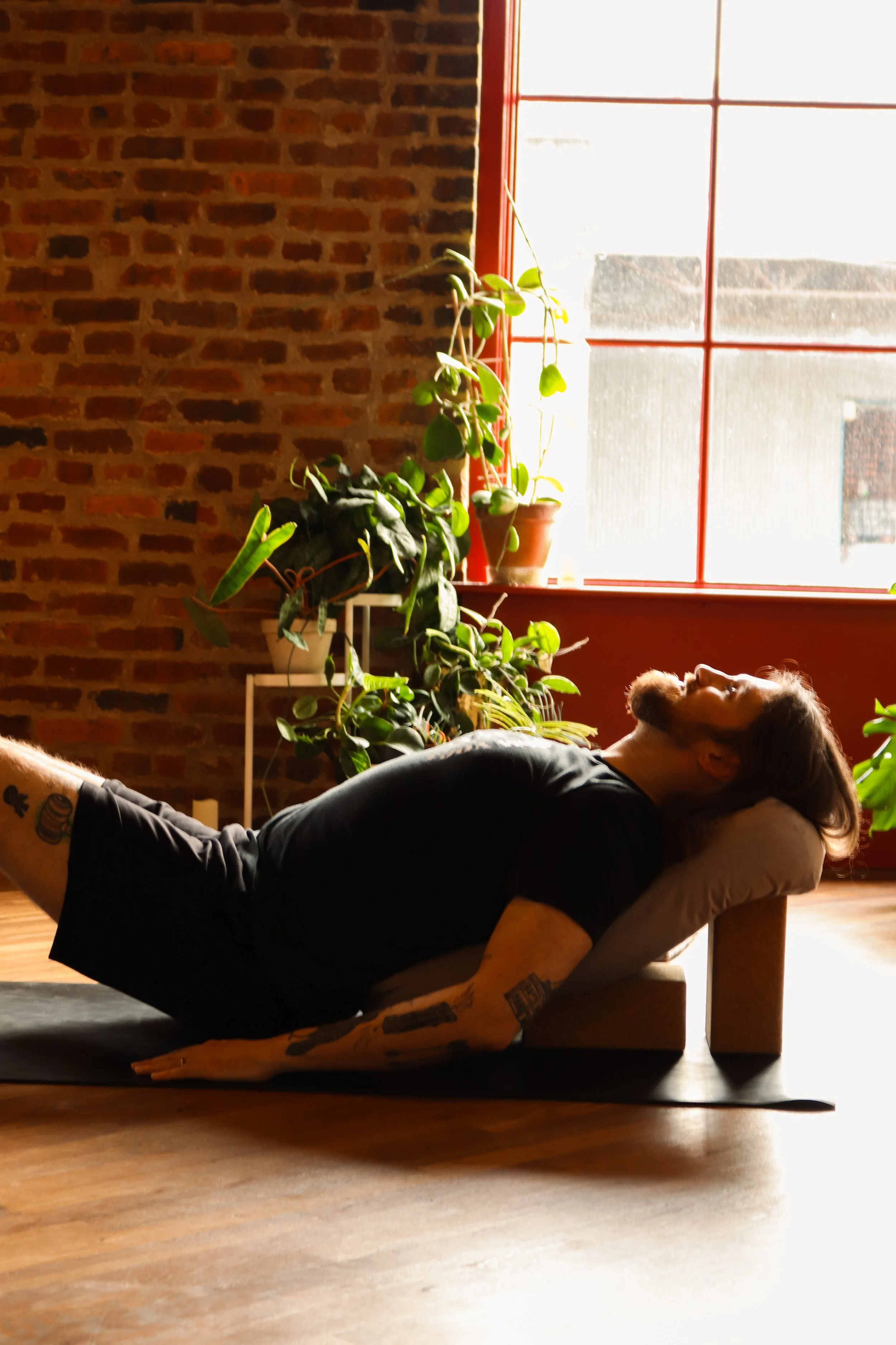 A person with tattoos lying on a yoga mat, performing a forearm plank yoga pose in a room with brick walls, a large window, and indoor plants.