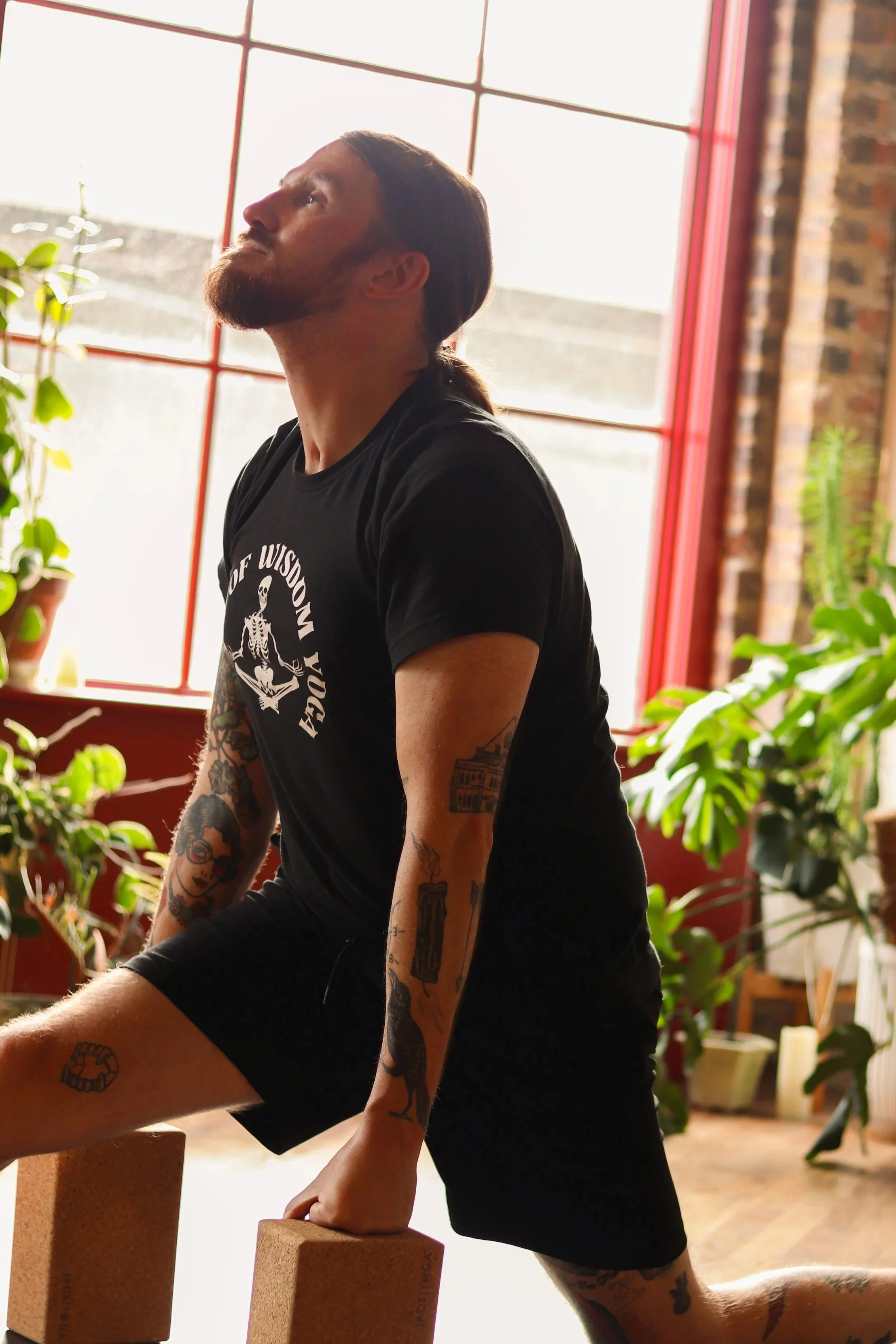 A man with tattoos and a beard stretching using yoga blocks in a room with large windows and plants.