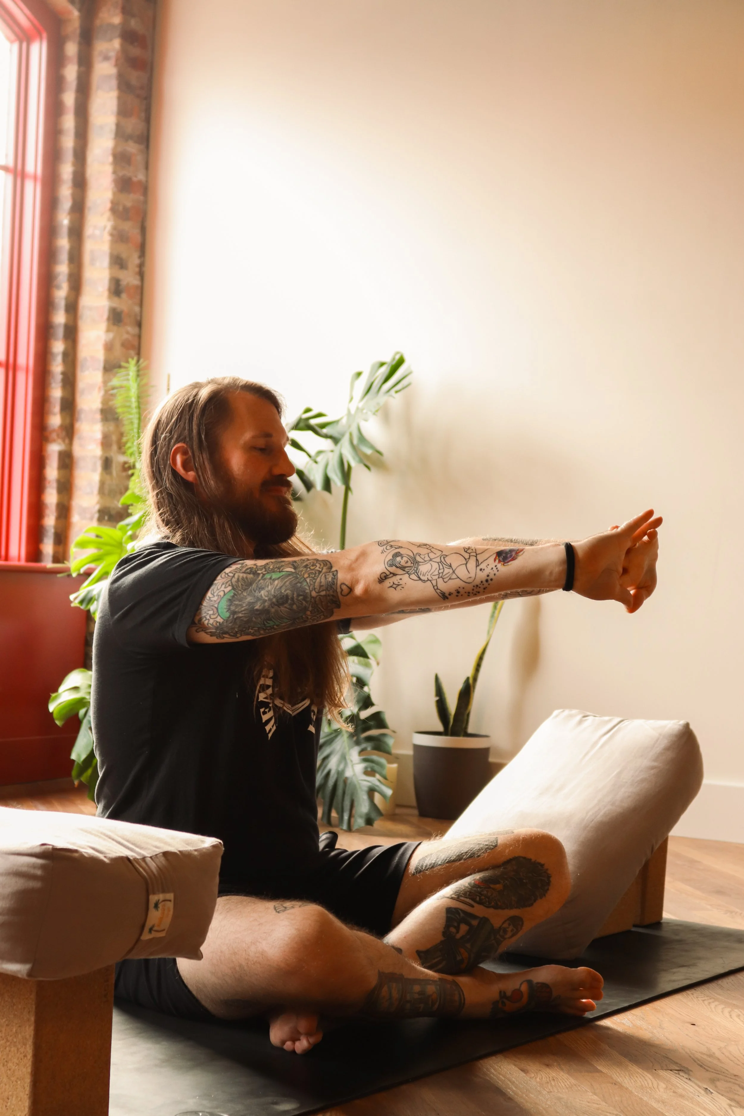 A man with long hair and tattoos is practicing yoga or meditation indoors, seated cross-legged on a yoga mat with his arms extended forward. There are plants in the background and a soft light coming through a window.