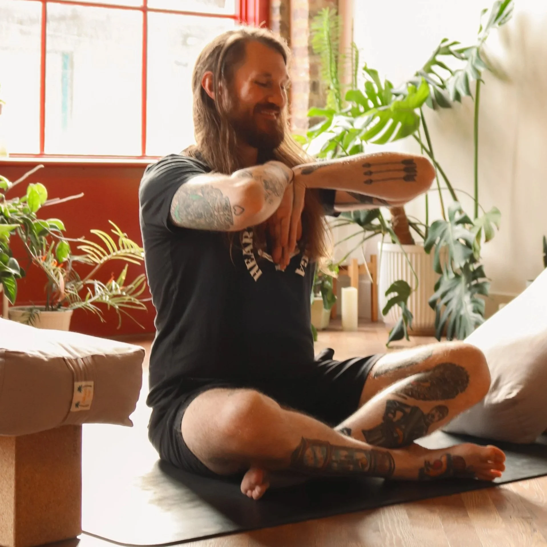 A man with long hair and a beard practicing yoga or meditation while seated on a cushion in a room filled with green plants, with a window and sunlight in the background.