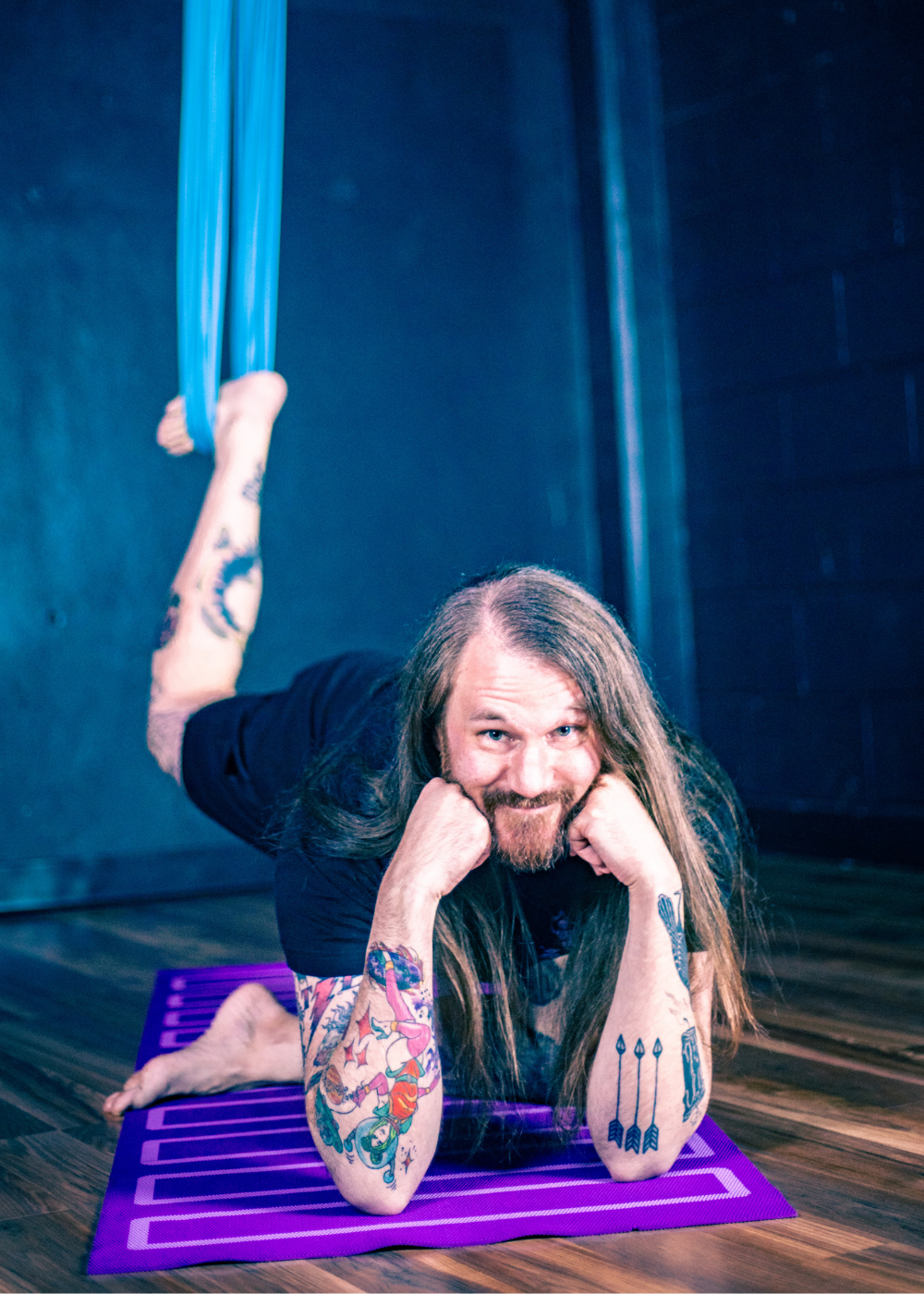 A man with long hair and tattoos practicing aerial yoga in a purple hammock and on a purple mat, resting on his elbows with his chin on his hands, smiling at the camera in a dimly lit room.