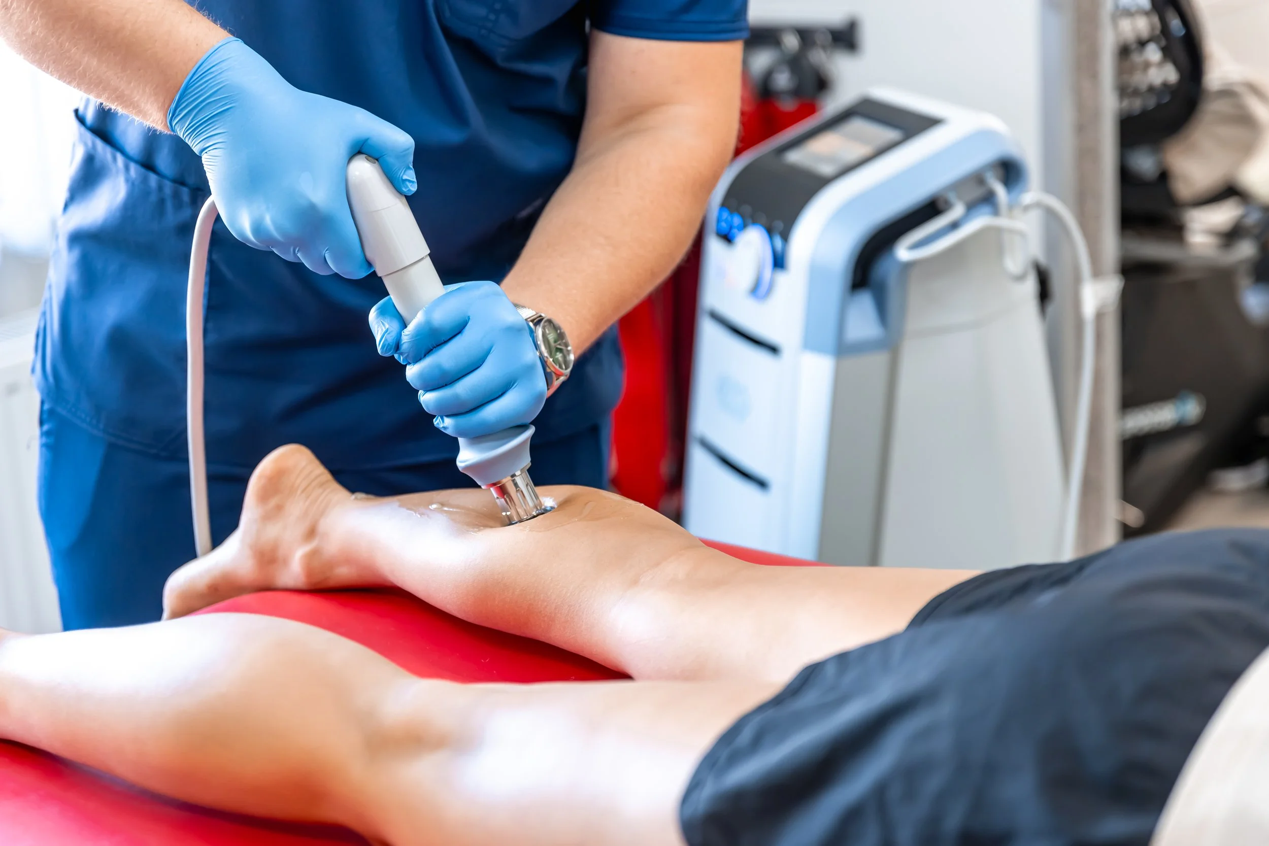 A chiropractor using shockwave therapy on a patient's leg in a clinical setting.