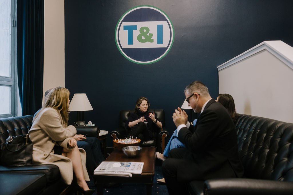 Group of people sitting in a meeting room with a T&I logo on the wall, chatting and listening.