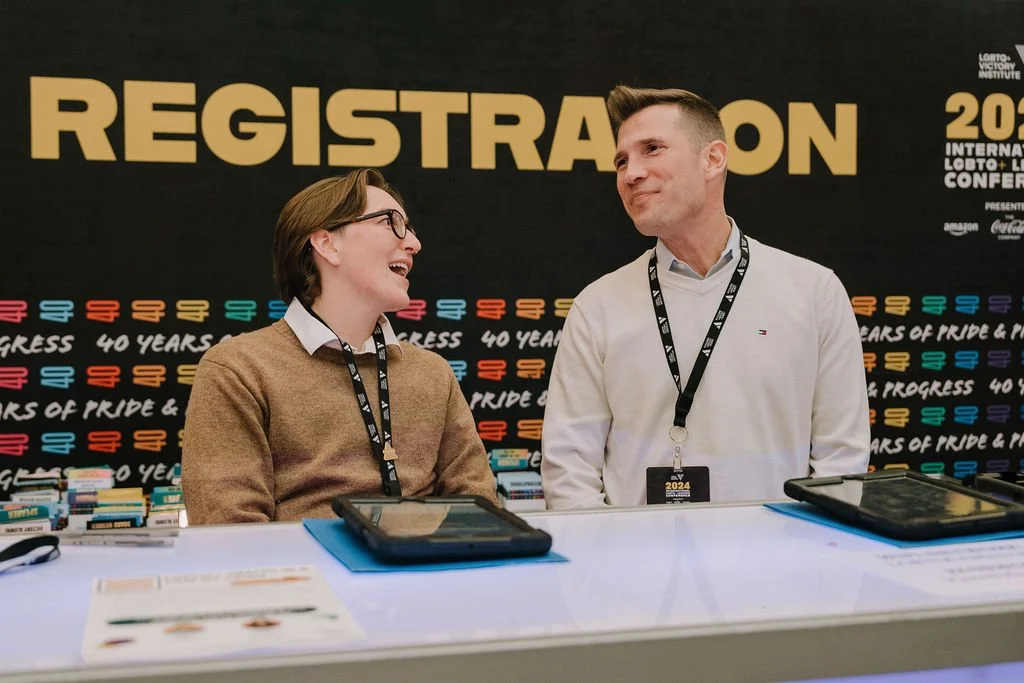 Two people with conference badges at registration desk, smiling and talking, behind a banner that says "REGISTRATION" against a black background with colorful text celebrating 40 years of pride and progress.