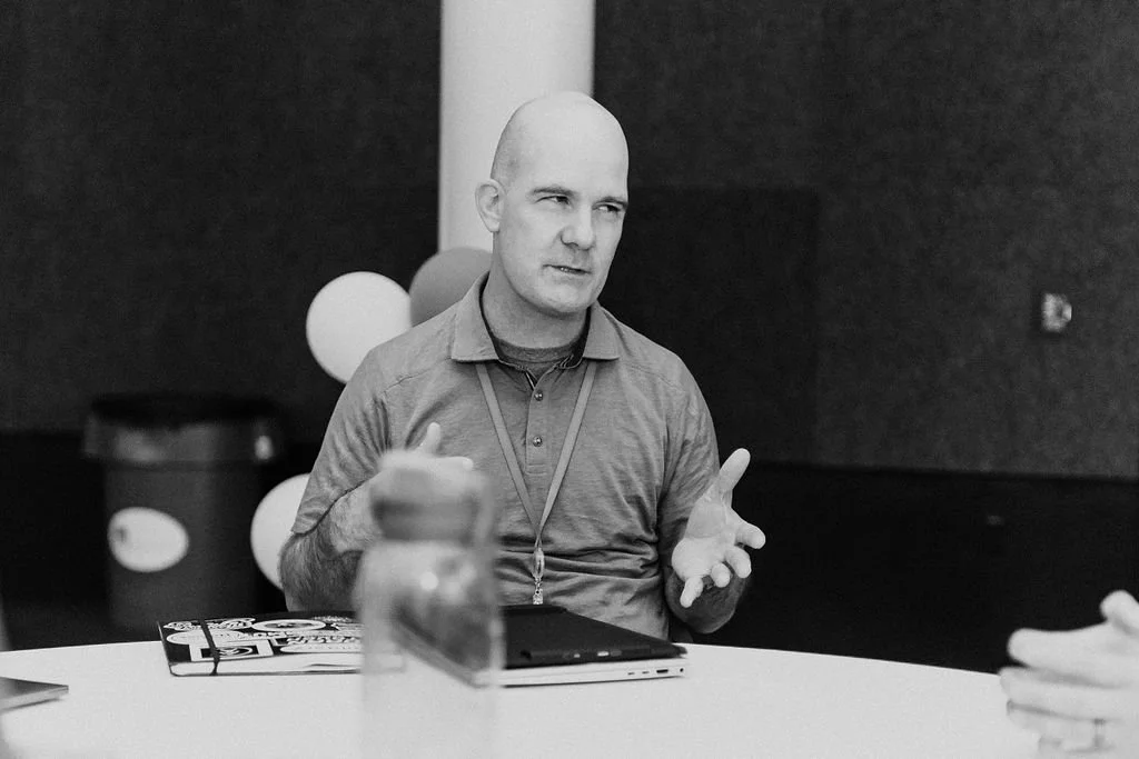 A man with a shaven head sitting at a table, gesturing with his right hand while talking. There are balloons in the background and a laptop and a magazine on the table.