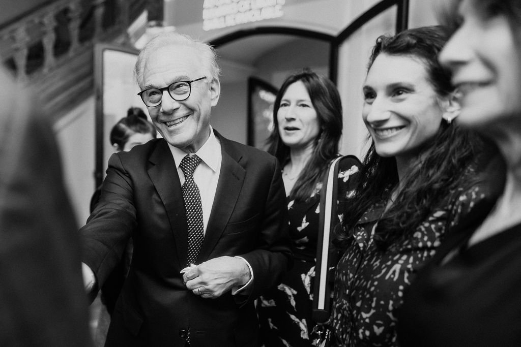 A group of people smiling and talking at an indoor event, with a man in a suit and glasses engaging in conversation.