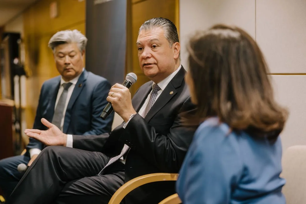 Three people engaged in a panel discussion or interview in a professional setting. The man in the center is holding a microphone, speaking, and gesturing with his hand. The person on the left is listening, and the person on the right, with back facin